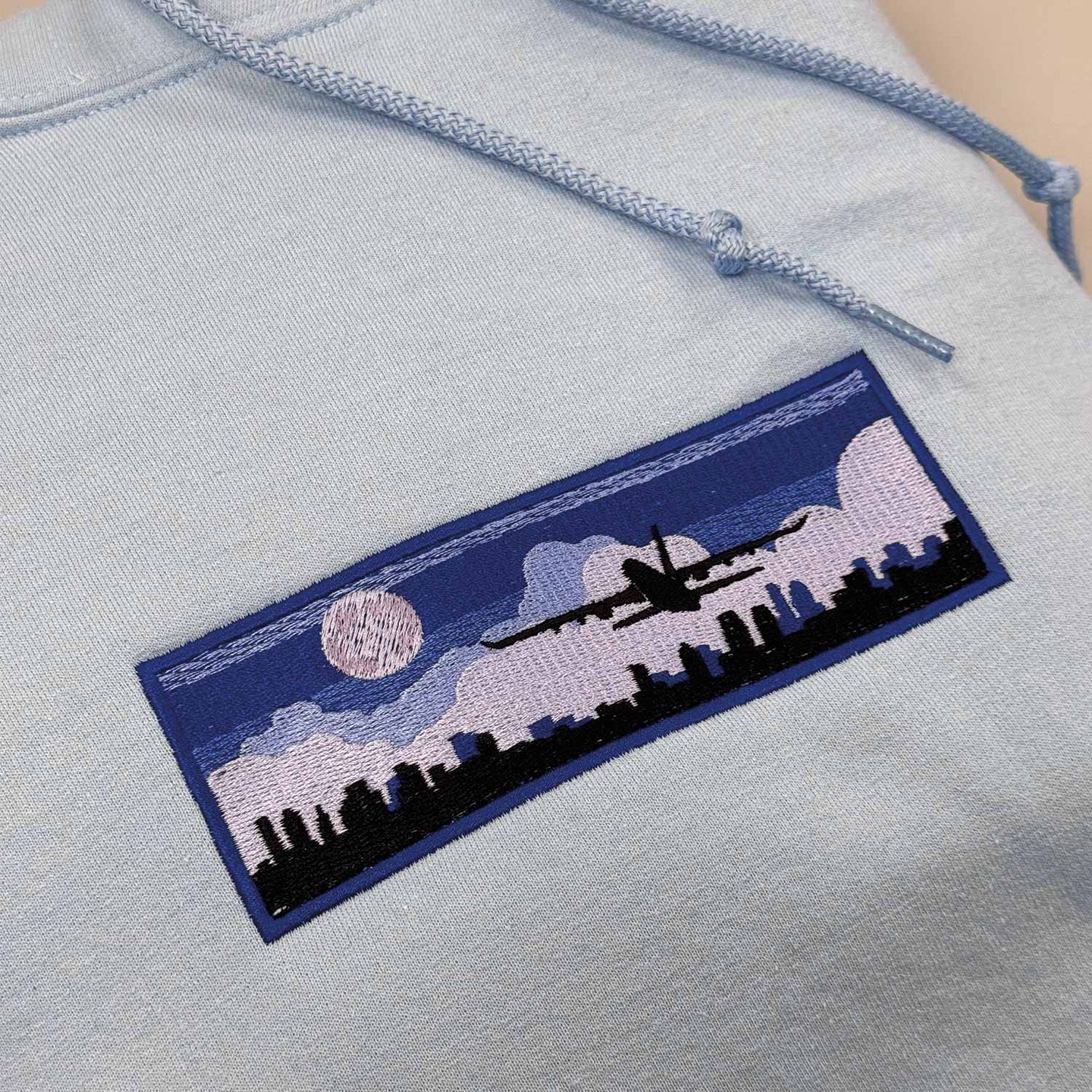 photo d'un plan rapproché d'un coton ouaté à capuchon bleu pâle avec une broderie d'un décollage d'avion dans la nuit