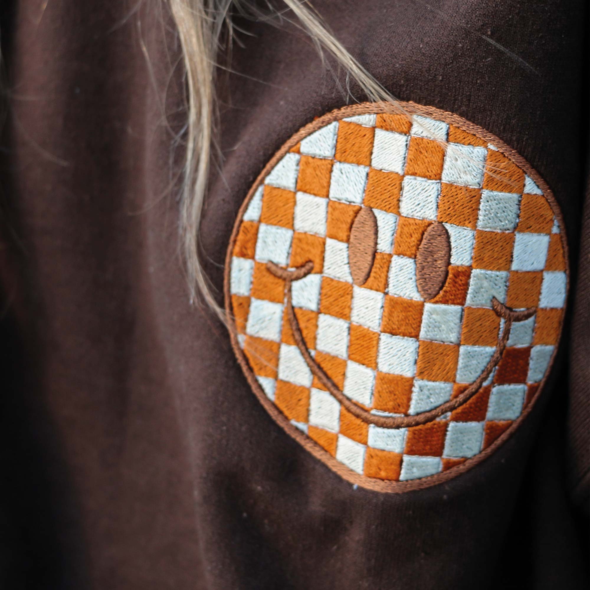 photo plan rapproché d'un crewneck brun avec une broderie de bonhomme sourire à motif quadrillé orange brun et blanc cassé