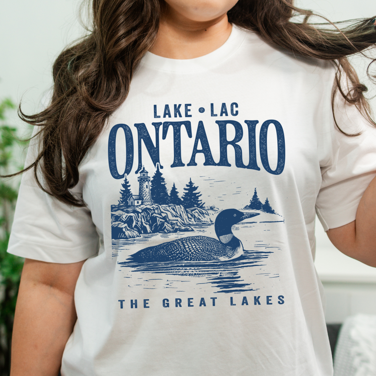 jeune femmes aux cheveux longs bruns frisés qui porte un t-shirt à manches courtes blanc ayant un imprimé du lac Ontario au Canada.
