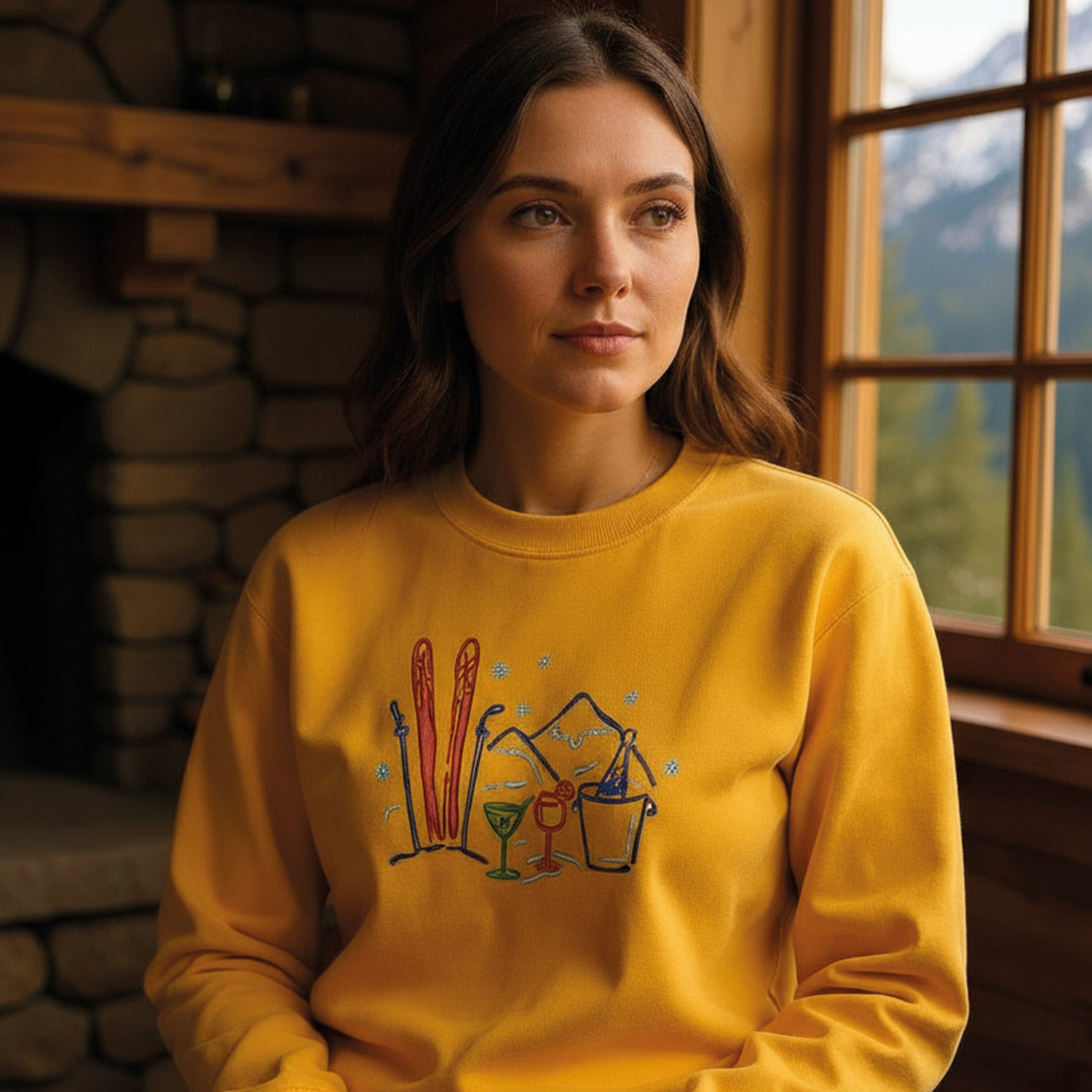 jeune femme aux cheveux bruns portant un coton ouaté à col rond jaune ayant une broderie d'illustrations dessinées à la main colorés de ski et de cocktails