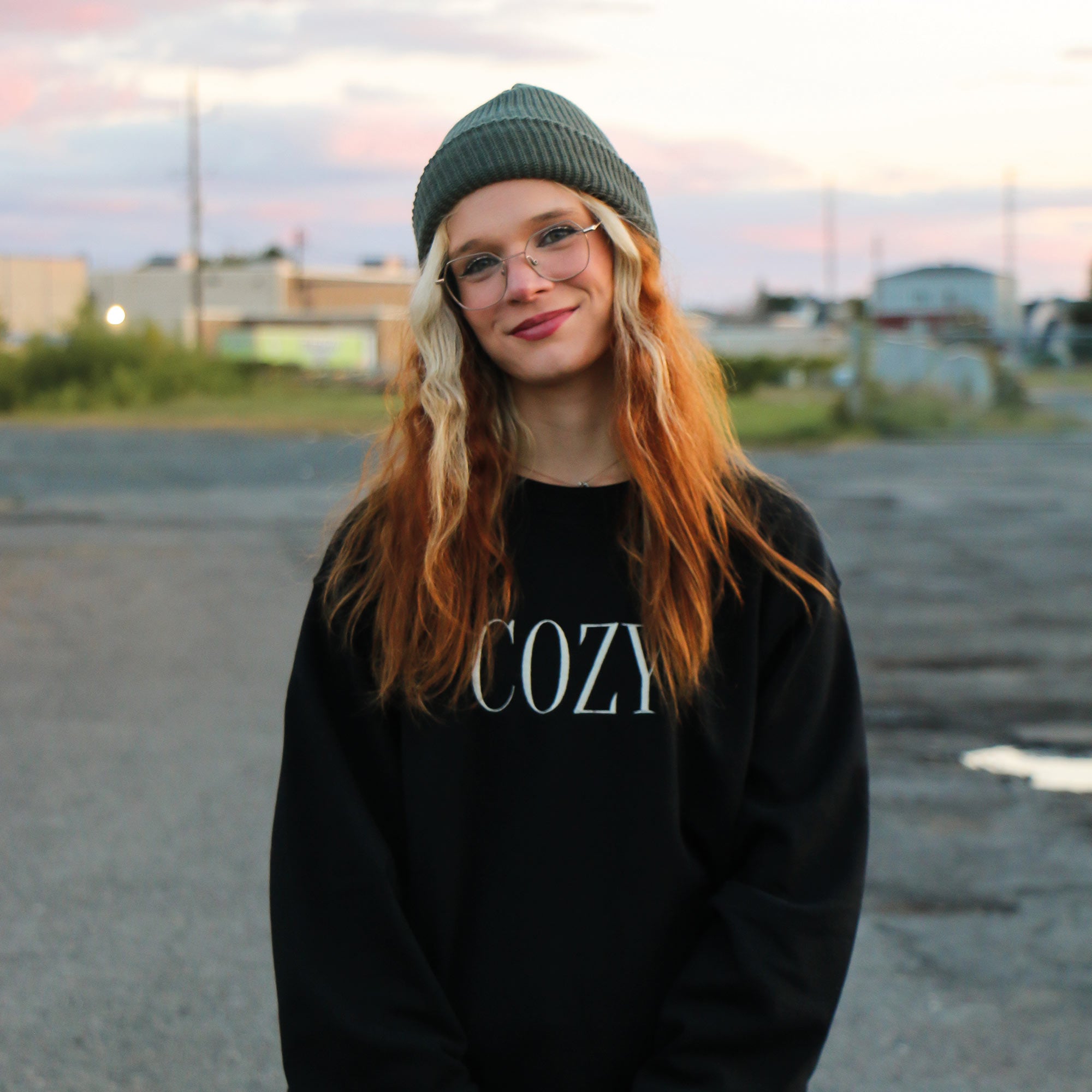 photo d'une jeune femme rousse portant un crewneck noir avec le mot COZY brodé en blanc crème au centre. Elle porte une tuque verte forêt