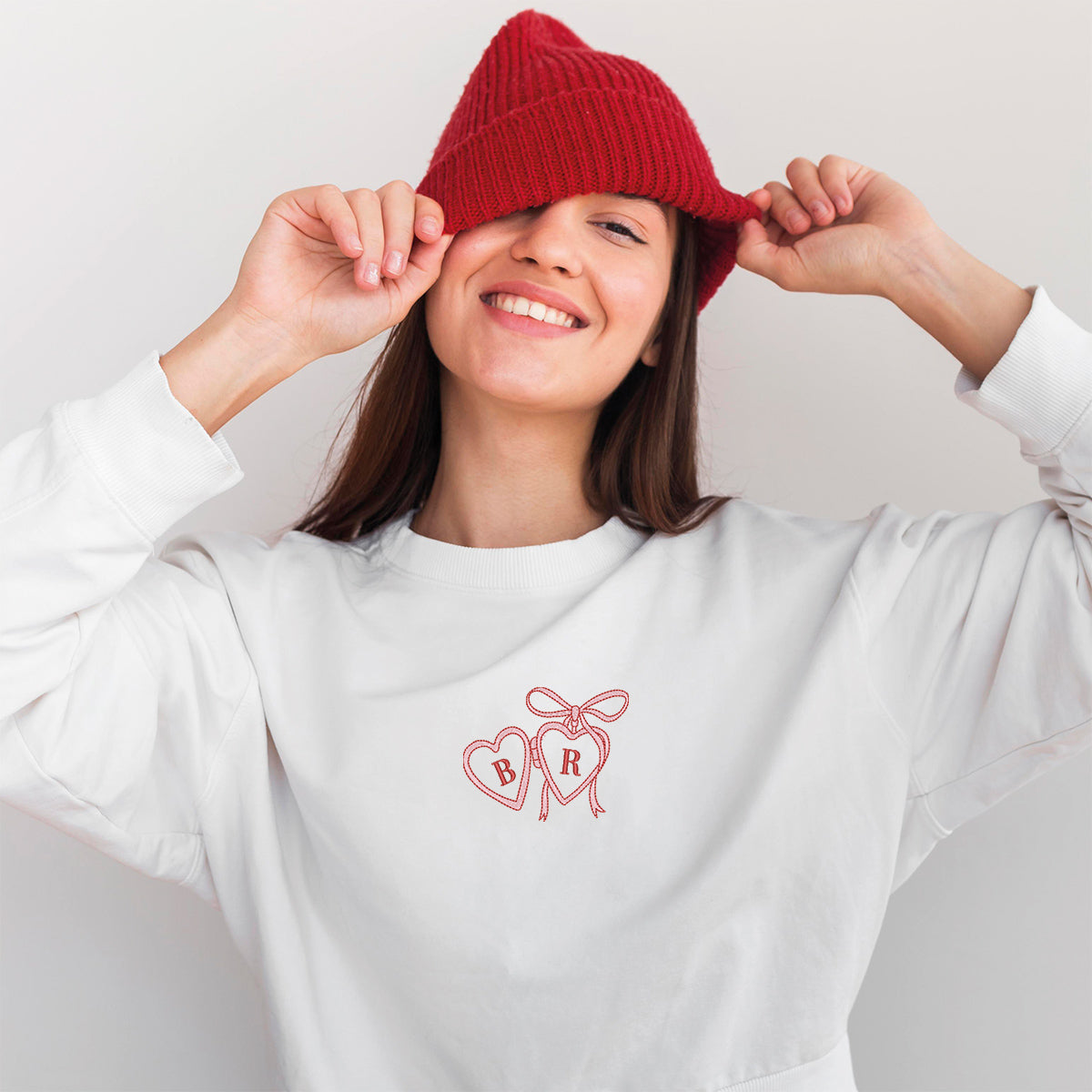 photo lifestyle d'une jeune femme brune portant une tuque rouge et un crewneck blanc avec une broderie d'un pendentif en forme de coeur rouge et rose centré avec des initiales à l'intérieur