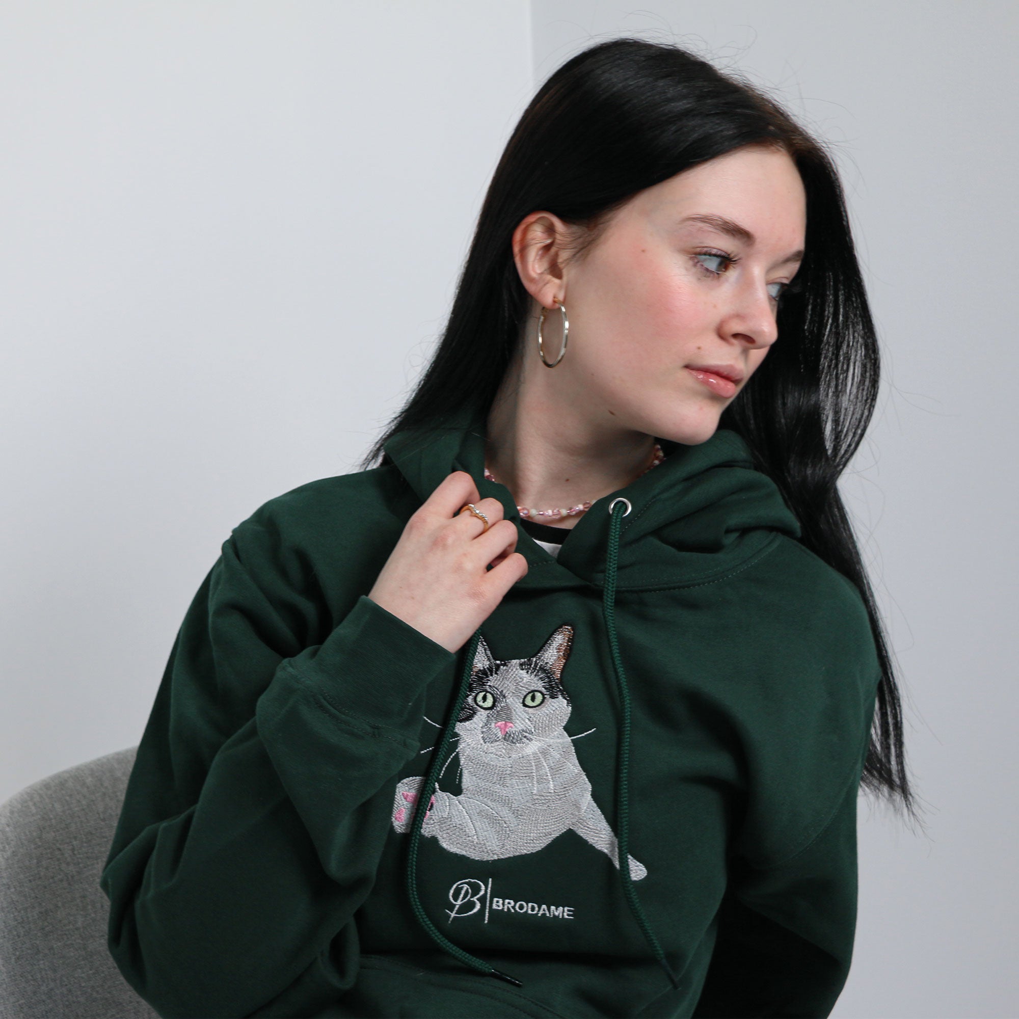 photo dans un studio d'une jeune femme aux cheveux bruns foncés portant un coton ouaté à capuchon vert forêt avec une broderie de son chat de type réaliste