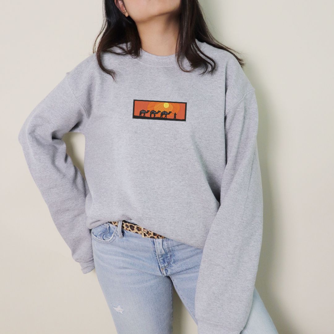 photo lifestyle d'une jeune femme portant un crewneck gris pâle avec une broderie de coucher de soleil dans le desert avec des chameaux.