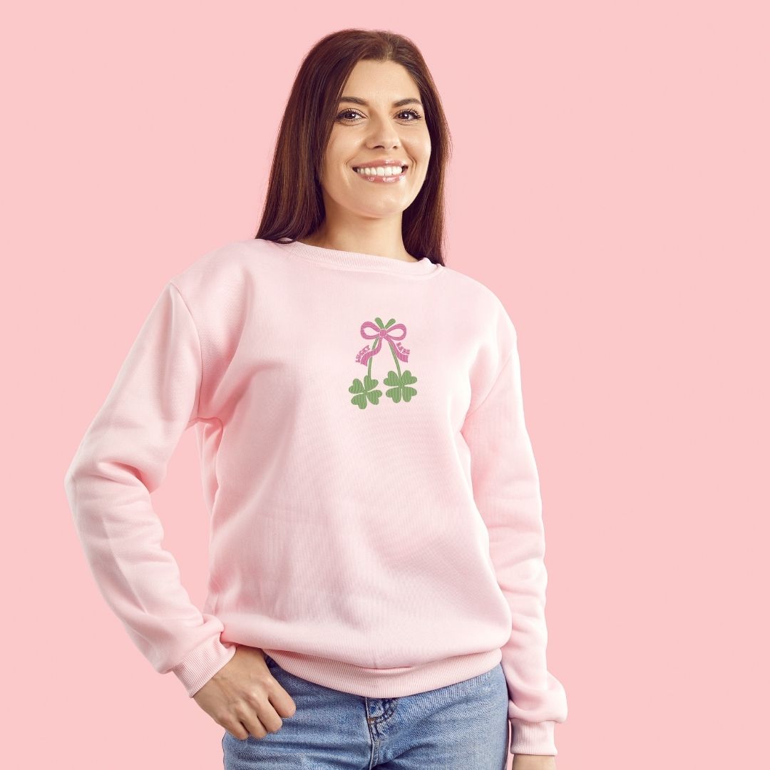 photo lifestyle d'une jeune femme aux cheveux brun portant un crewneck rose pâle avec broderie de trèfles à 4 feuilles et une boucle rose. photo en studio sur fond rose pâle