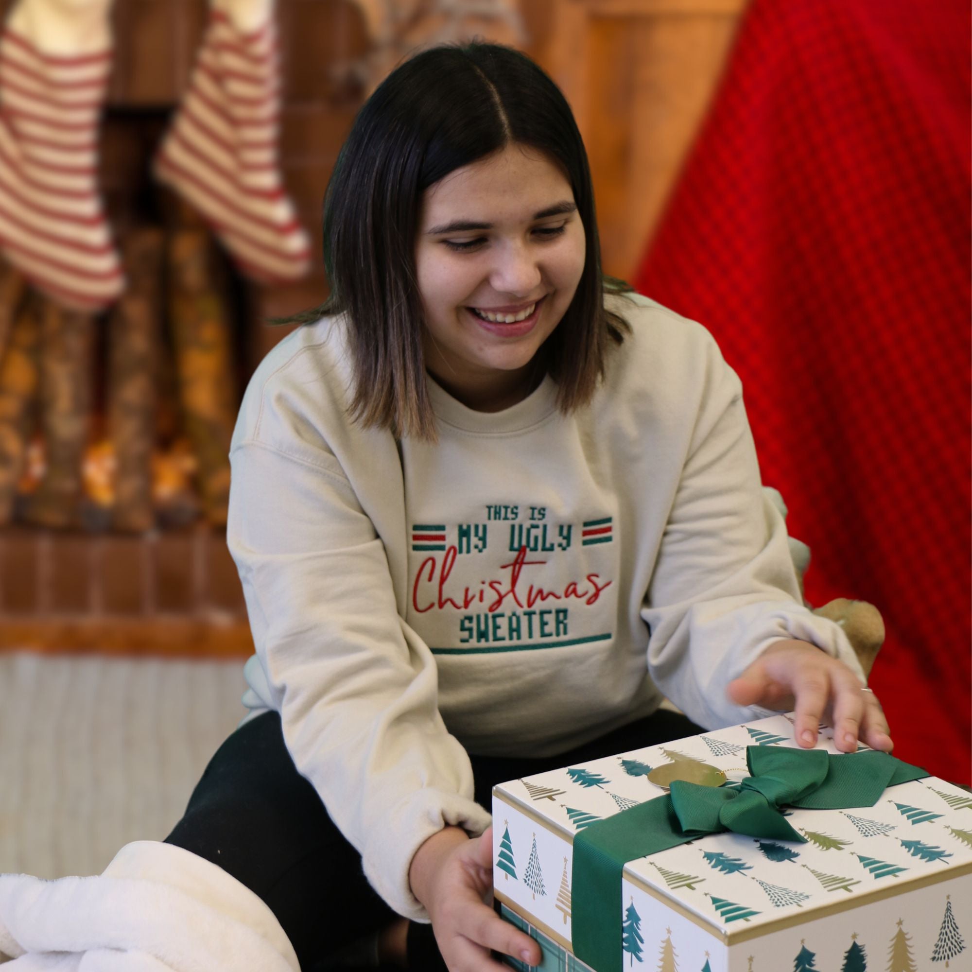photo d'un jeune femme brune portant un crewneck beige avec une broderie this is my ugly christmas sweater en vert et rouge
