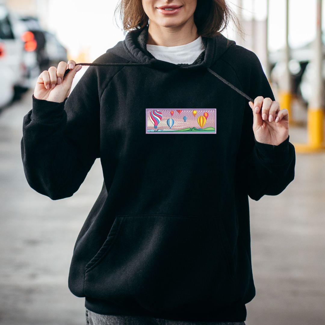 photo lifestyle d'un jeune femme brune portant un coton ouaté à capuchon noir avec une broderie complexe de plusieurs montgolfières au centre dans un stationnement