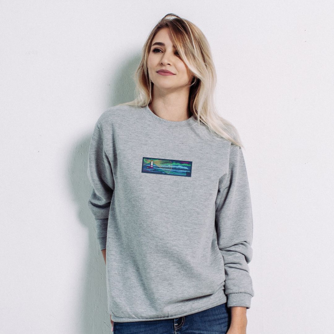 photo lifestyle d'une jeune femme blonde portant un crewneck gris pâle brodé d'un phare sur le bord de la mer en pleine nuit