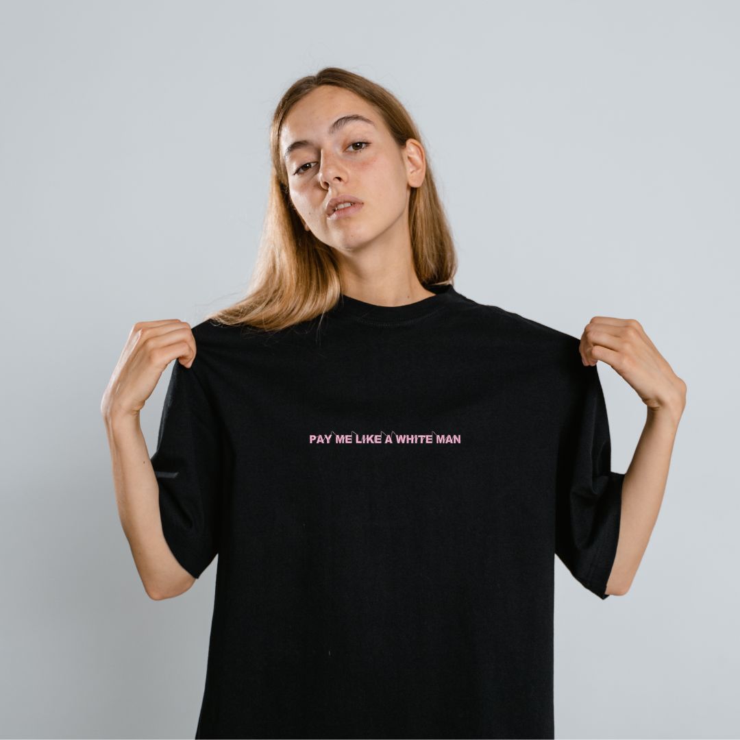photo d'une jeune femme blonde portant un t-shirt à manche courtes noir brodé "pay me like a white man" en rose pâle