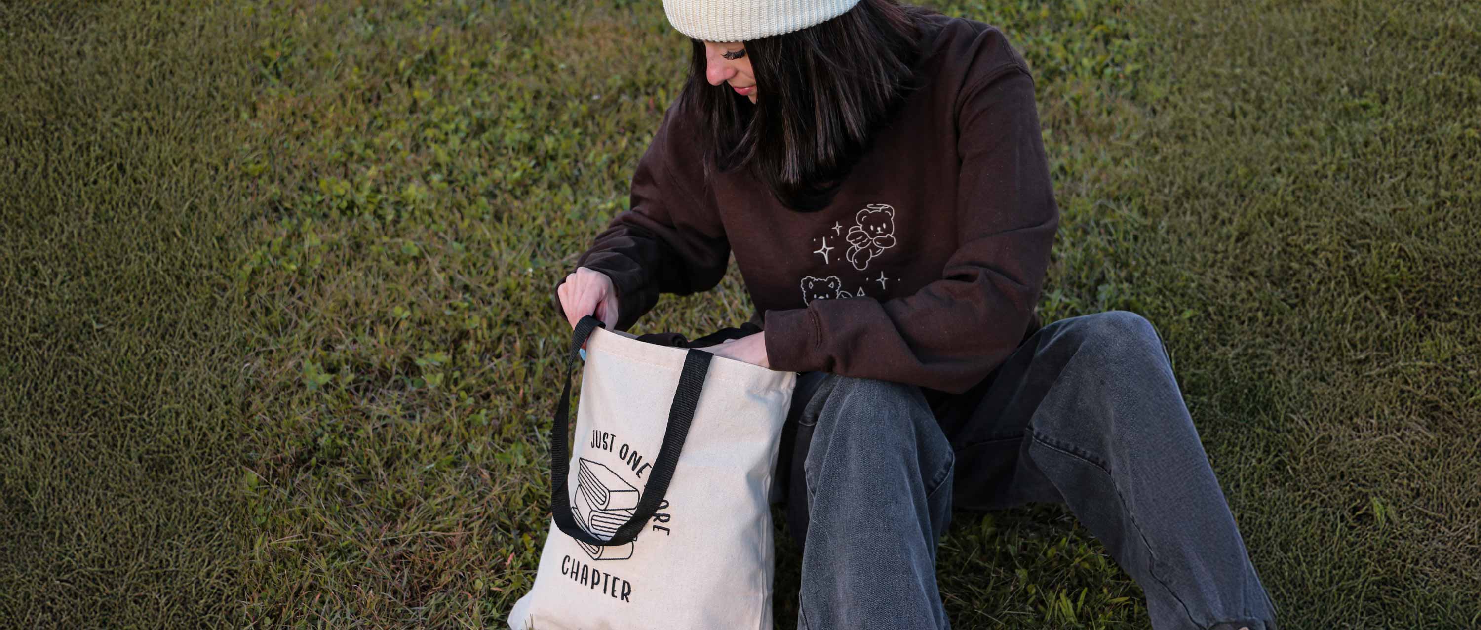 jeune femme assise dans la pelouse portant un crewneck brun brodé avec des petits oursons, une tuque blanche et un sac fourre-tout beige et noir avec des livres brodés dessus