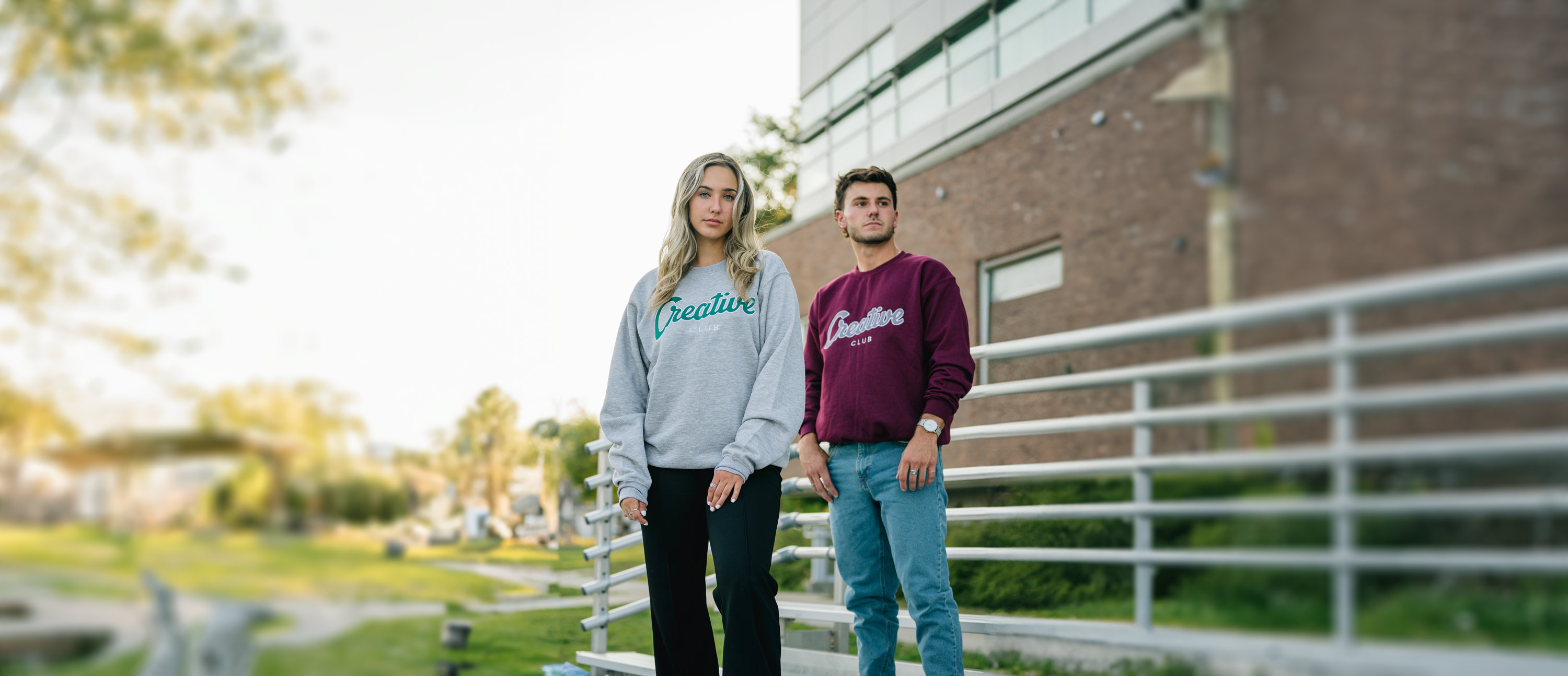 photo agrandie d'une jeune femme et d'un jeune homme portant le crewneck avec une broderie creative club au centre. Ils se situent dans un estrade près d'un bâtiment