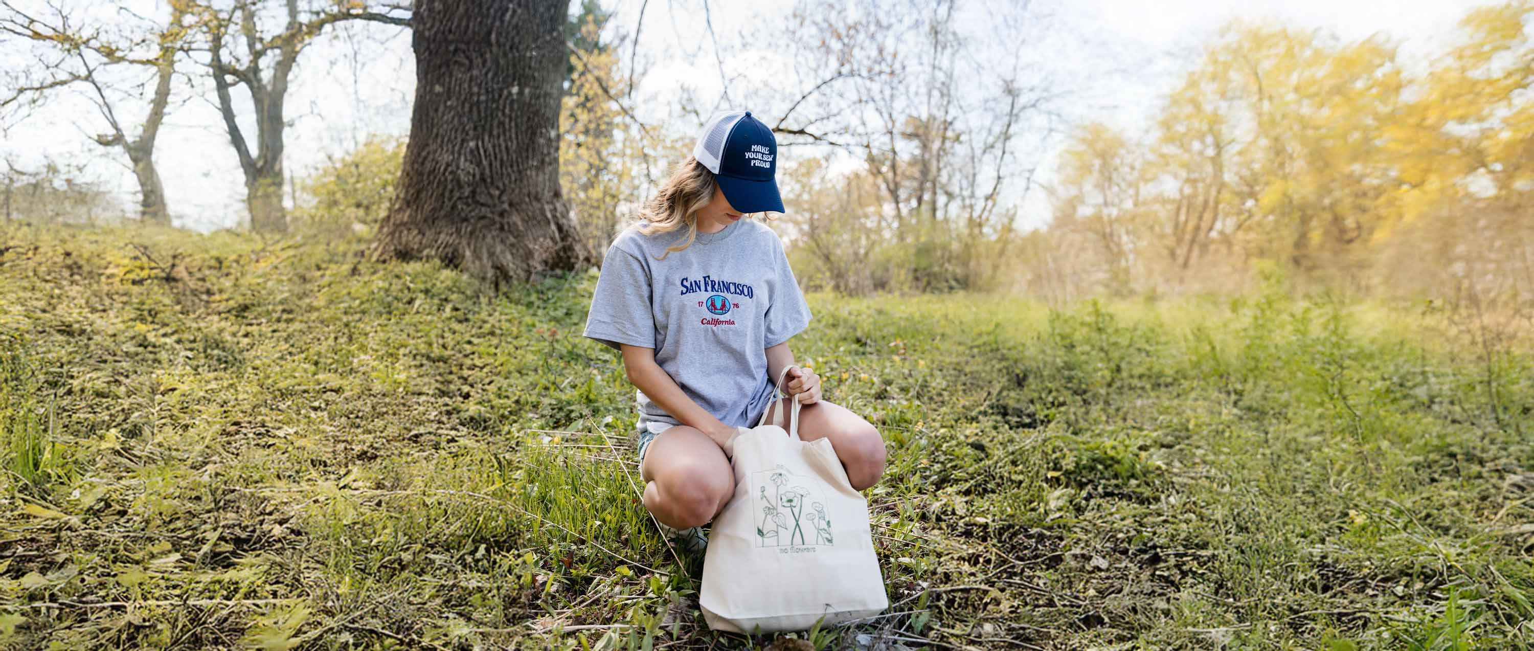 photo d'une jeune fille dans un petit boisé portant un t-shirt gris une casquette bleu marine et blanche et un tote bag brodés venant de chez brodame