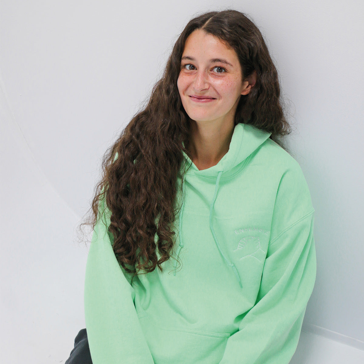 jeune femme aux cheveux longs bruns portant un coton ouaté à capuchon menthe avec une broderie de poignée de main et l'écriture "Ensemble" au-dessus, en arche, en blanc