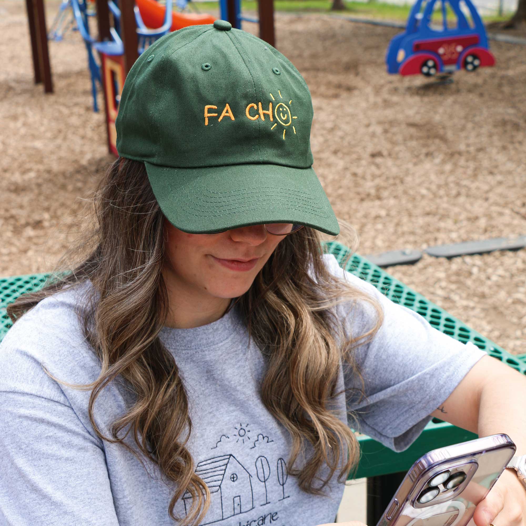 jeune femme dans un parc pour enfants portant une casquette vert foret avec une broderie "fa cho" avec un petit soleil qui remplace le "o". 