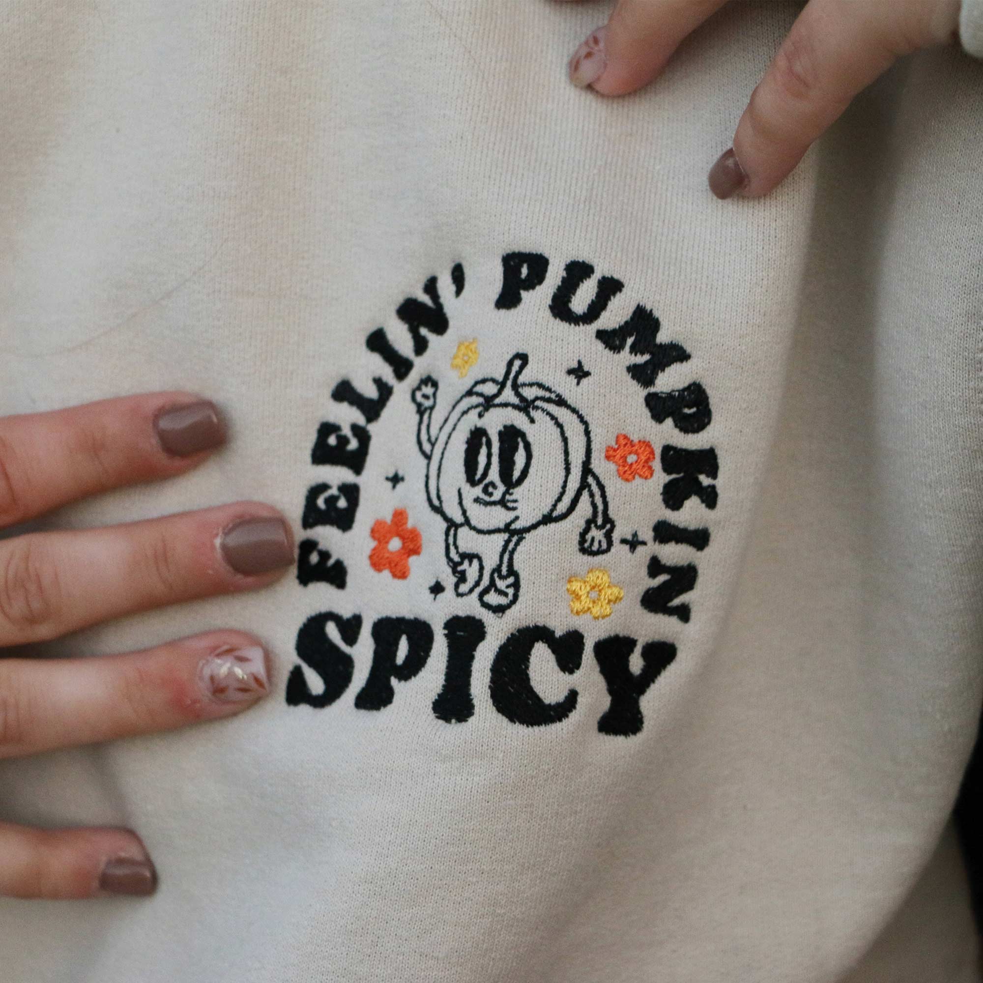 photo de plan rapproché d'une broderie d'automne citrouille animée pumpkin spicy avec des petites fleurs oranges et jaunes sur un crewneck beige