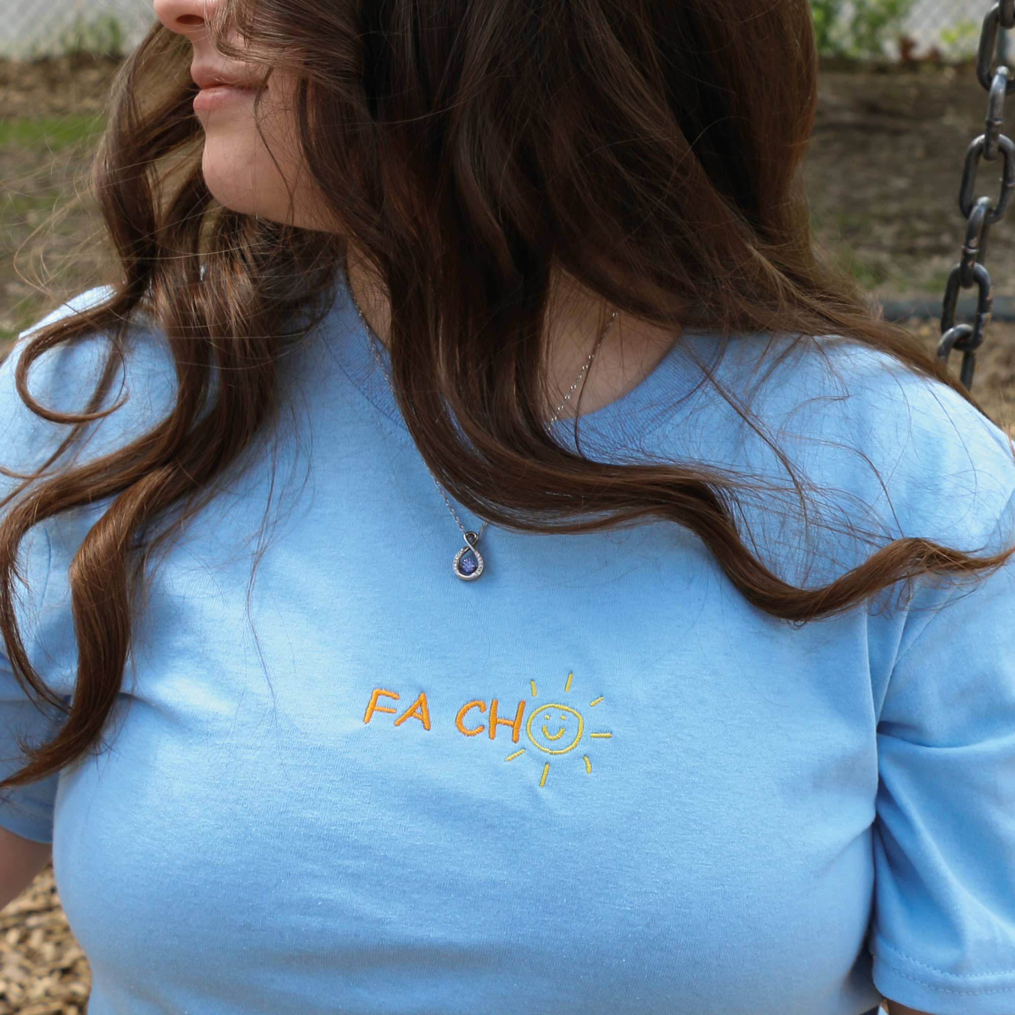plan rapproché d'un t-shirt à manches courte bleu pâle avec une broderie "fa cho" orange et jaune brodé au centre du t-shirt. Il est porté par une jeune femme aux cheveux bruns bouclés