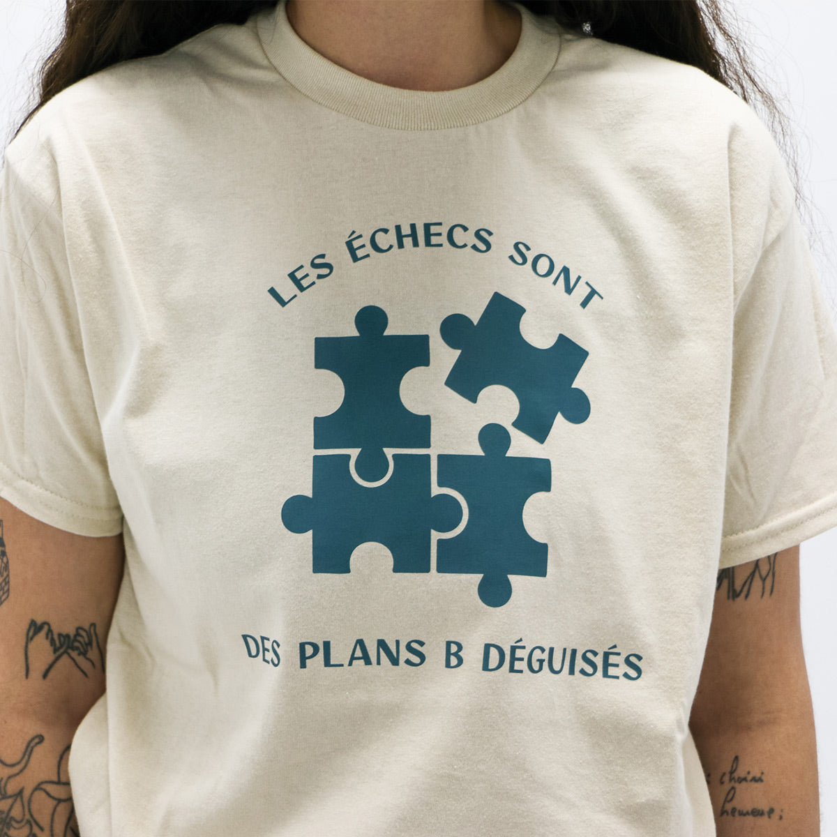 plan rapproché du t-shirt beige avec un imprimé "les échecs sont des plans B déguisés" vert foncé, avec des morceaux de casse-tête. porté par une jeune femme tatouée