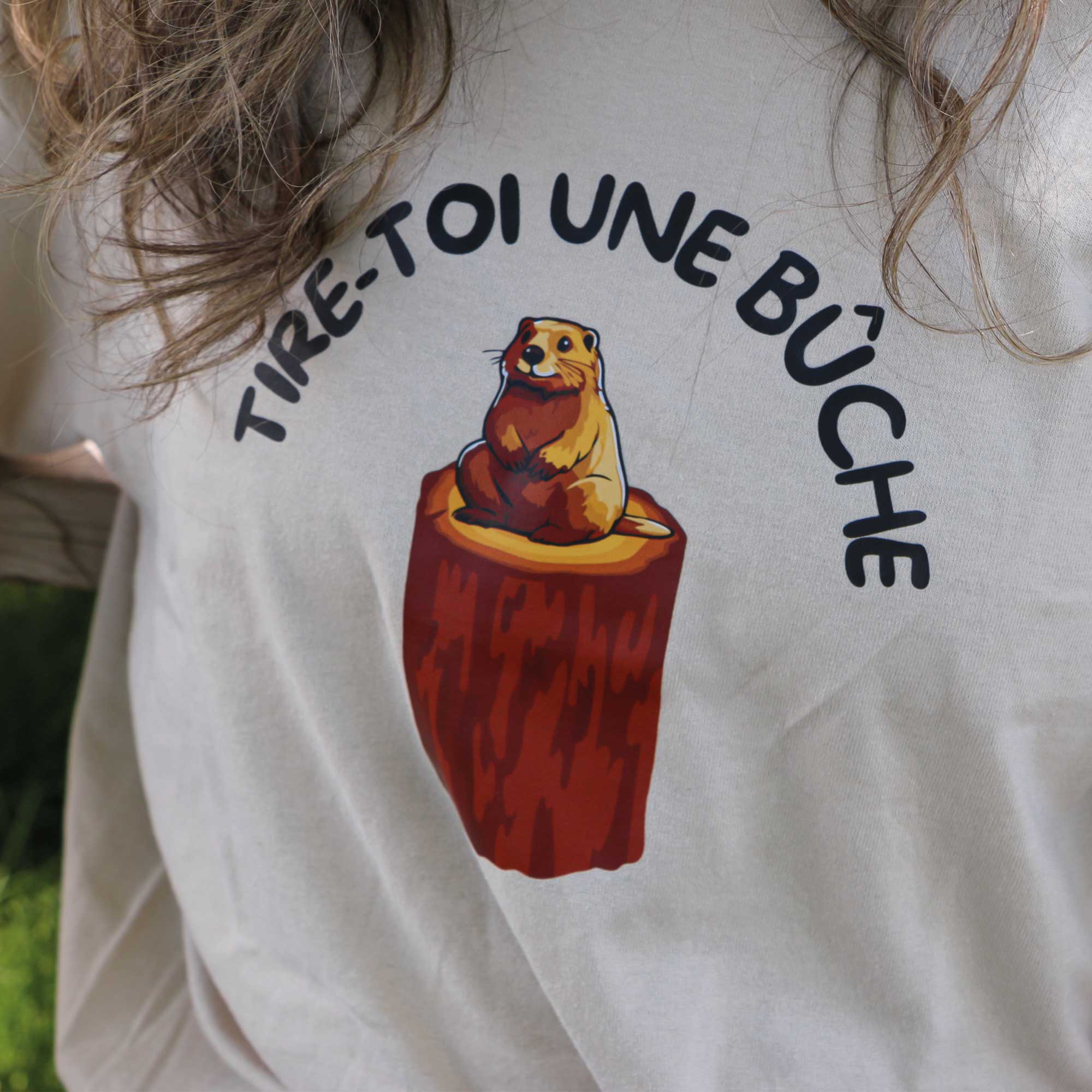 plan rapproché d'un t-shirt beige avec un imprimé d'un castor sur une buche avec l'inscription "tire-toi une bûche" en arc. Expression québécoise