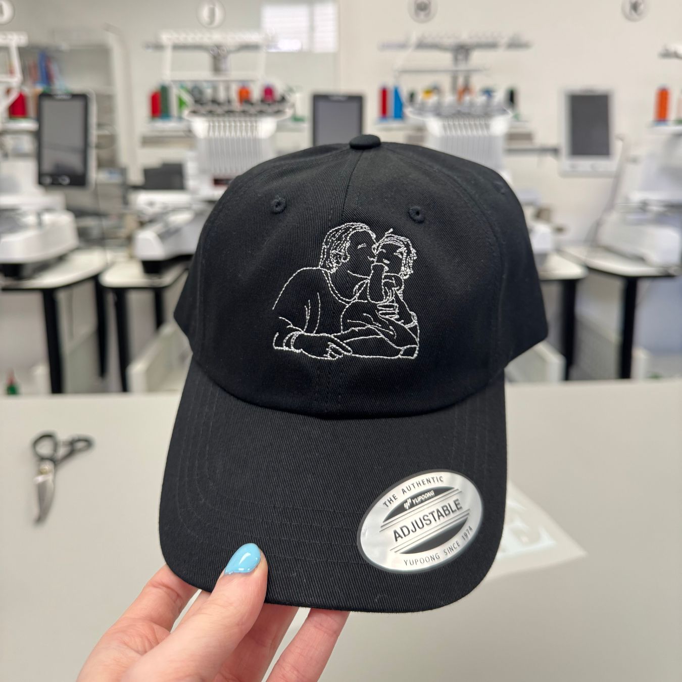 plan rapproché d'une casquette noire avec une silhouette brodée en blanc faite à partir d'une photo. Il y a des brodeuses en arrière. La casquette vient tout juste de se faire broder.