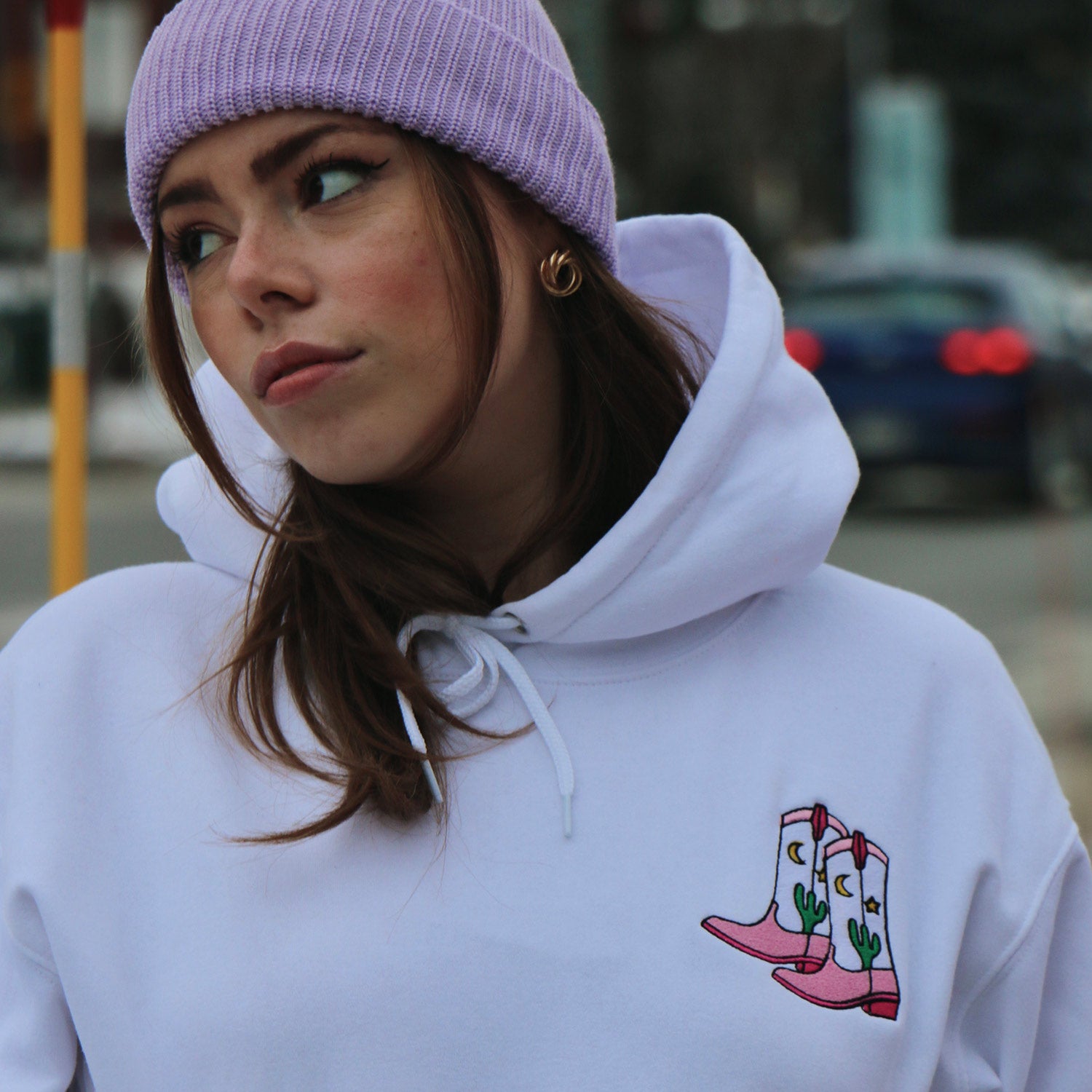 photo d'une jeune femme avec les cheveux bruns portant une tuque lilas et un hoodie blanc avec des bottes de cowboy. Illustration par Oska, broderie par Brodame