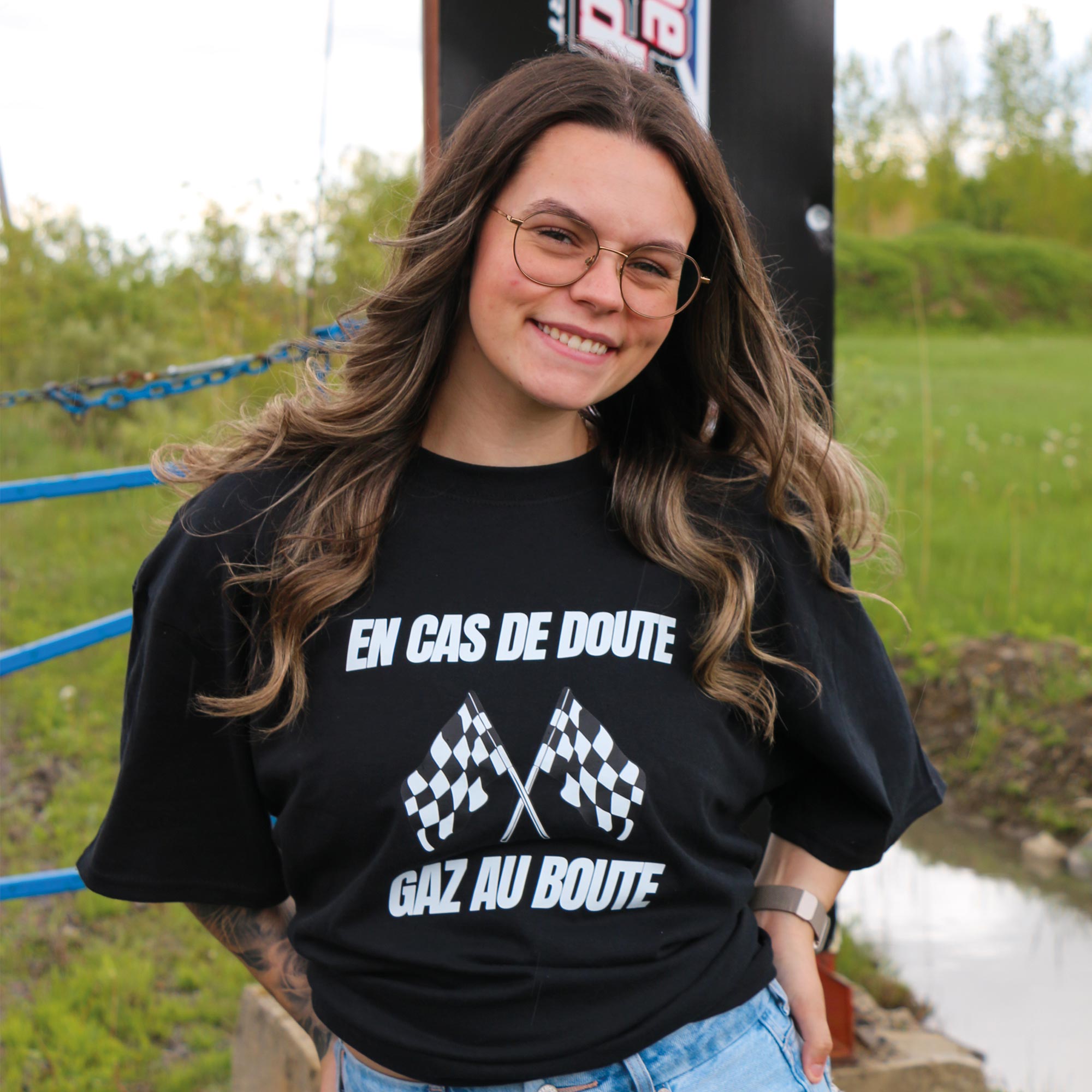 photo lifestyle d'une jeune femme souriante aux cheveux bruns bouclés portant un t-shirt noir "en cas de doute, gaz au boute" avec des drapeaux de course