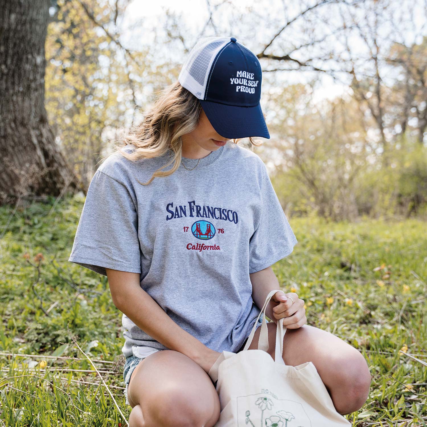 jeune femme portant une casquette bleu marine et blanche avec la broderie make yourself proud et un t-shirt gris pâle san francisco