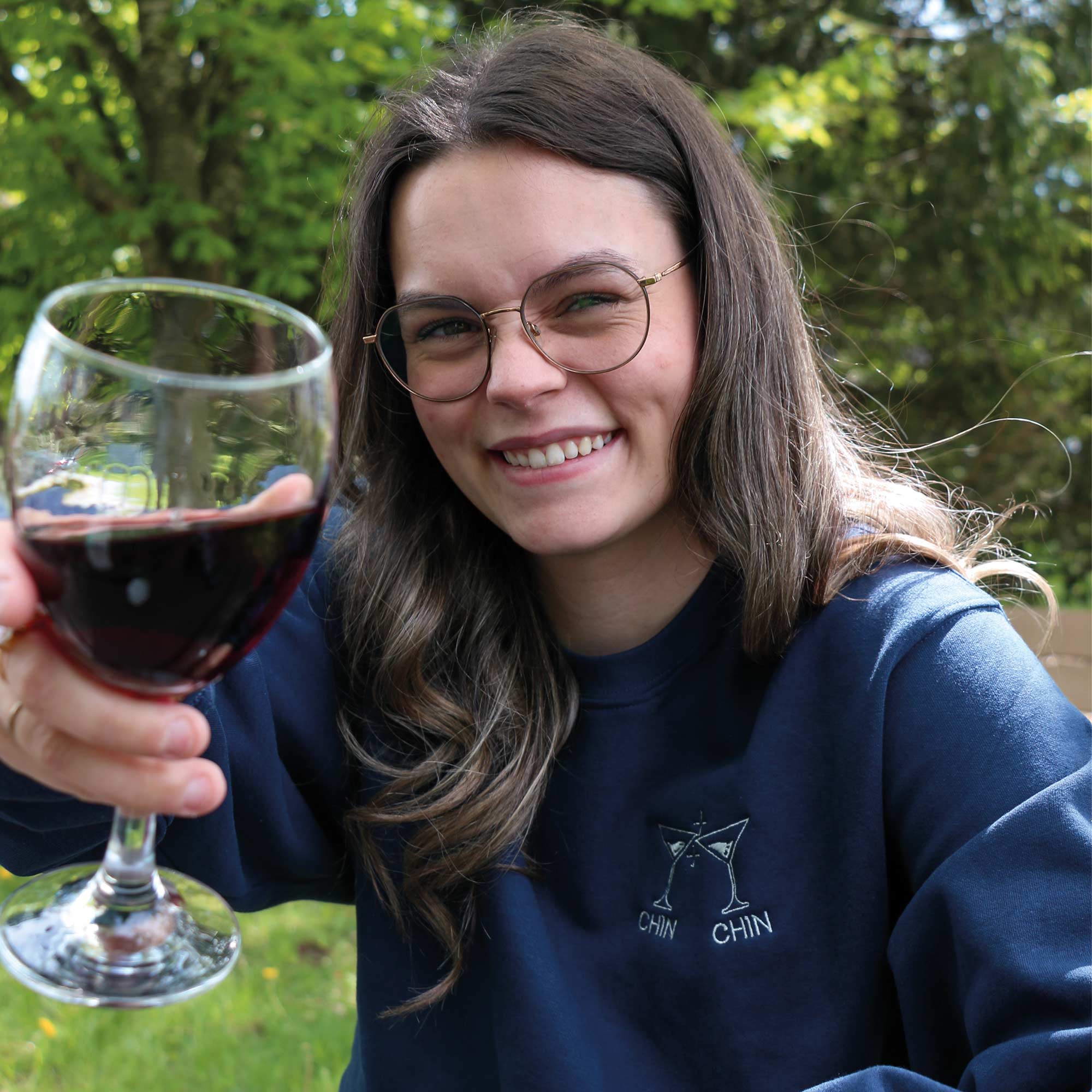 photo lifestyle d'une jeune femme souriante portant un crewneck bleu marin avec une broderie de verre de cocktail et l'écriture "chin chin" en dessous en blanc. elle a une coupe de vin dans ses mains