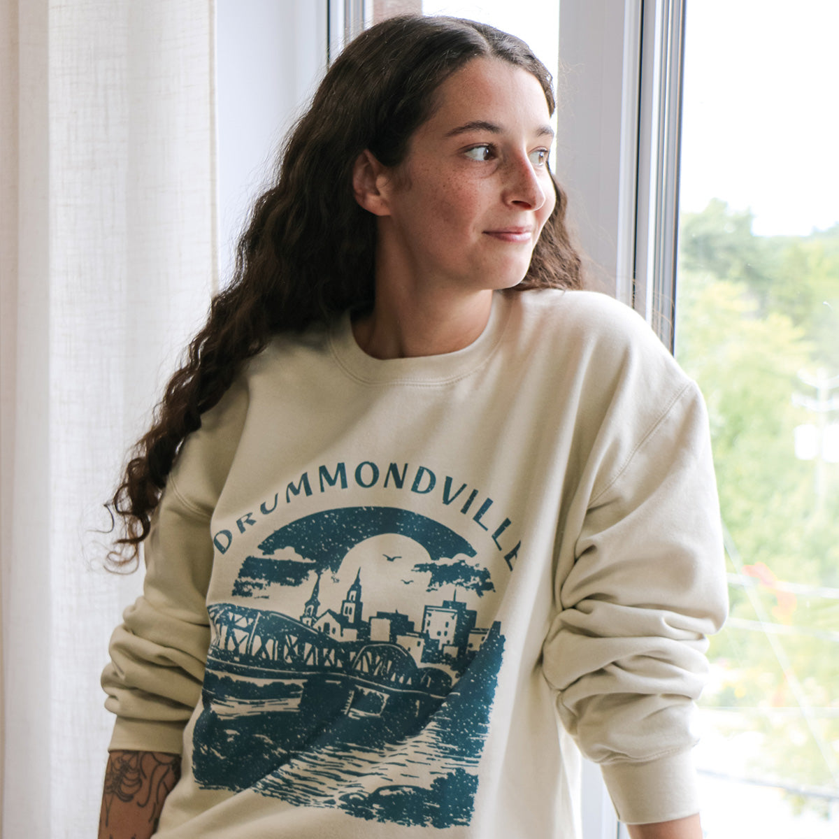 photo d'une jeune femme aux cheveux bruns frisés portant un crewneck beige avec une impression "Drummondville" avec une image du pont en turquoise foncé