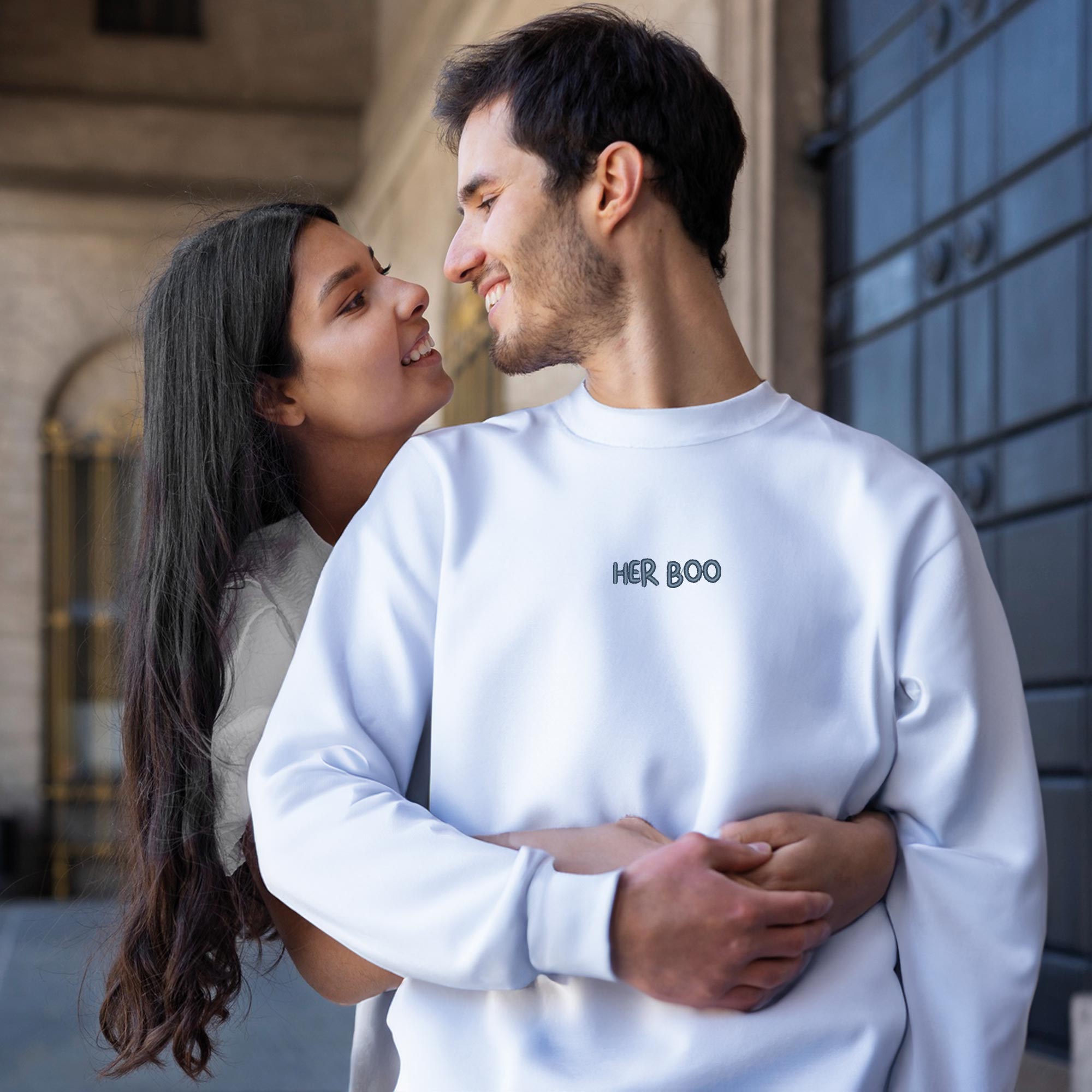 photo d'un couple s'enlançant. L'homme porte un sweatshirt blanc avec une broderie her boo bleu marin