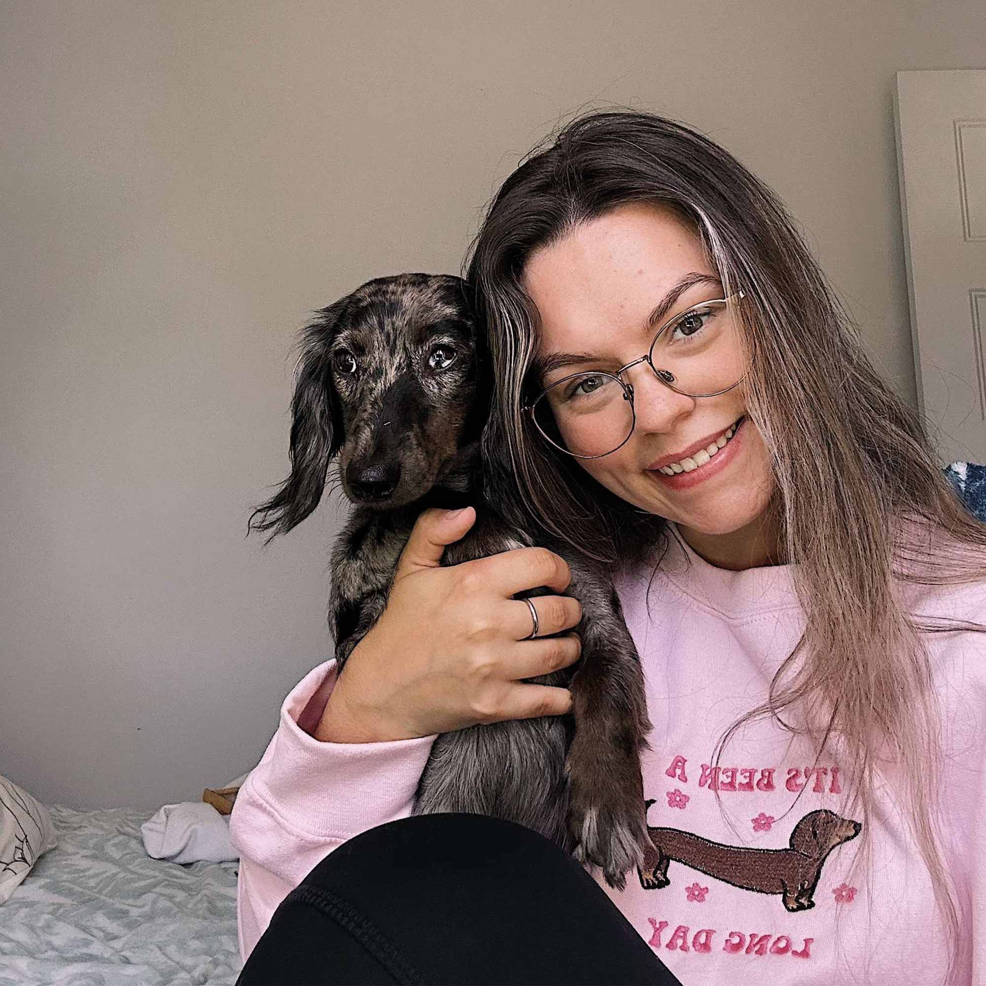 jeune femme aux cheveux bruns portant un crewneck rose pâle avec son chien saucisse teckel 