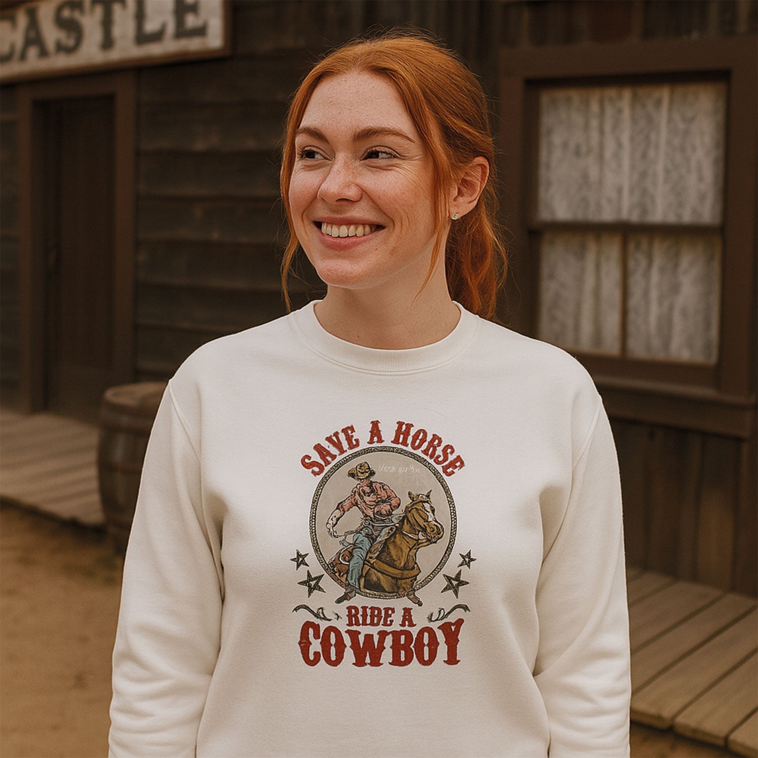 photo lifestyle d'une jeune femme aux cheveux roux posée devant un décor western. Elle porte un crewneck blanc avec un imprimé country et l'écriture "save a horse, ride a cowboy"