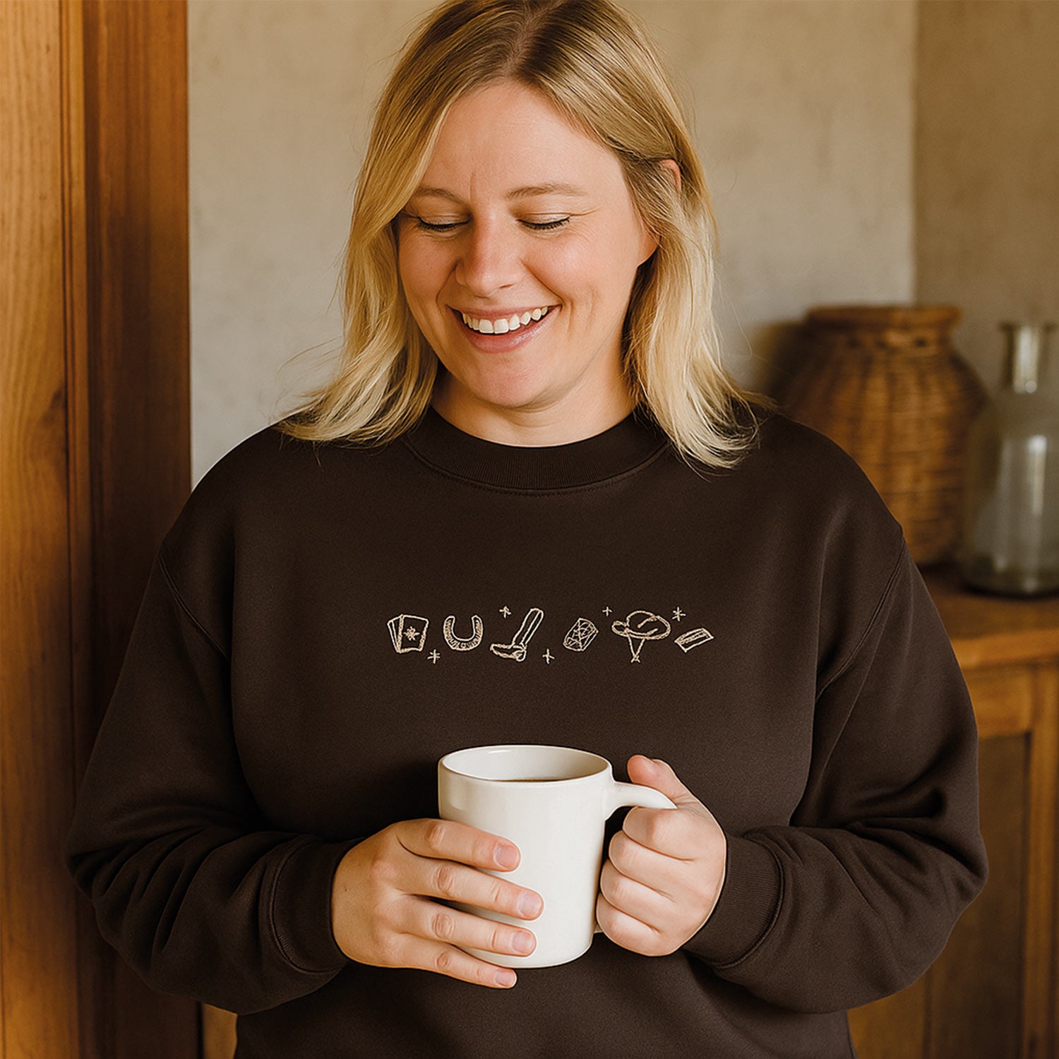 photo de style lifestyle d'une femme aux cheveux blonds souriant en portant un crewneck brun avec une broderie western beige et avant une tasse de café dans ses mains