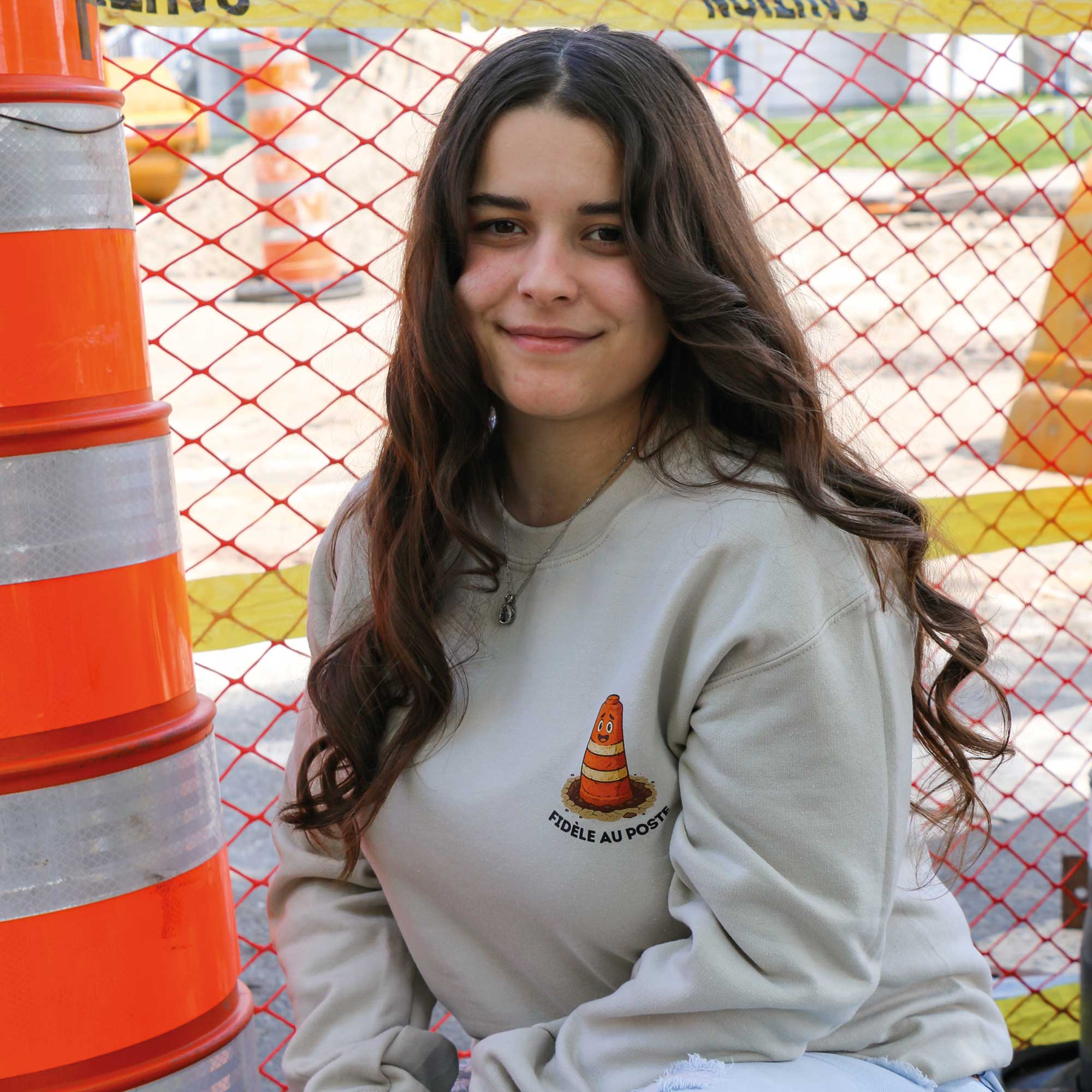 photo lifestyle d'une jeune femme aux cheveux bruns foncés dans une zone de construction portant un crewneck beige avec un imprimé de cône avec l'écriture "fidèle au poste" en dessous pour faire référence au routes du québéc