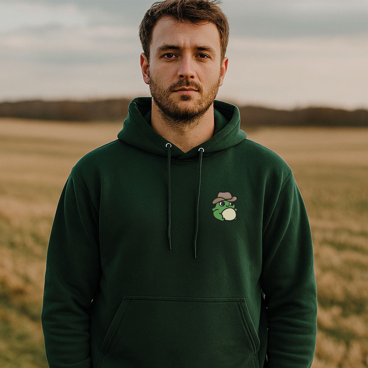 jeune homme portant un hoodie vert foret dans un champ de campagne avec une grenouille portant un chapeau de cowboy brodé au coeur