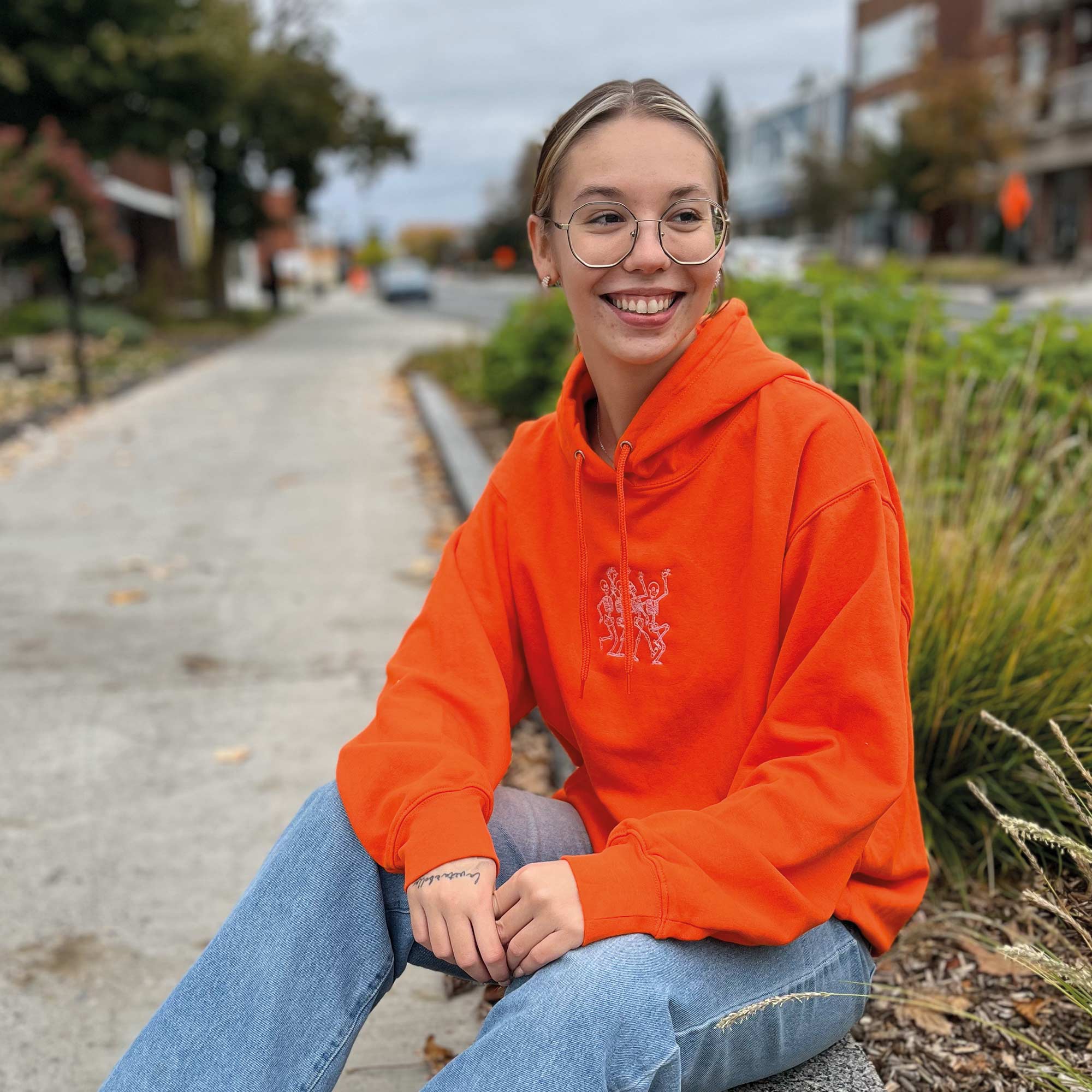 jeune femme souriante portant un coton ouaté à capuchon orange avec une broderie de squelettes qui dansent