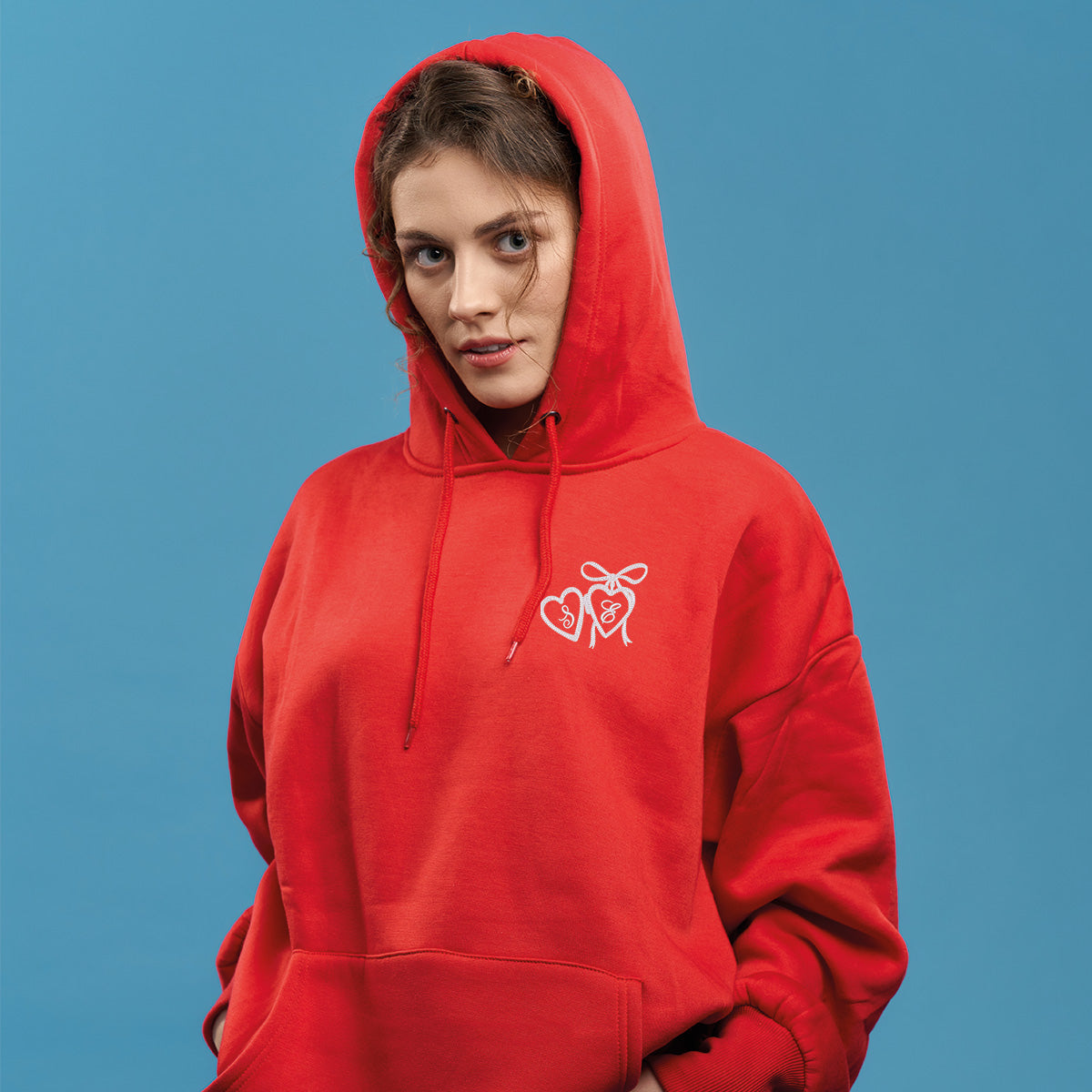 photo lifestyle d'une jeune femme aux cheveux bruns portant un coton ouaté à capuchon rouge avec une broderie de pendentif en forme de coeur blanc ayant des initiales à l'intérieur