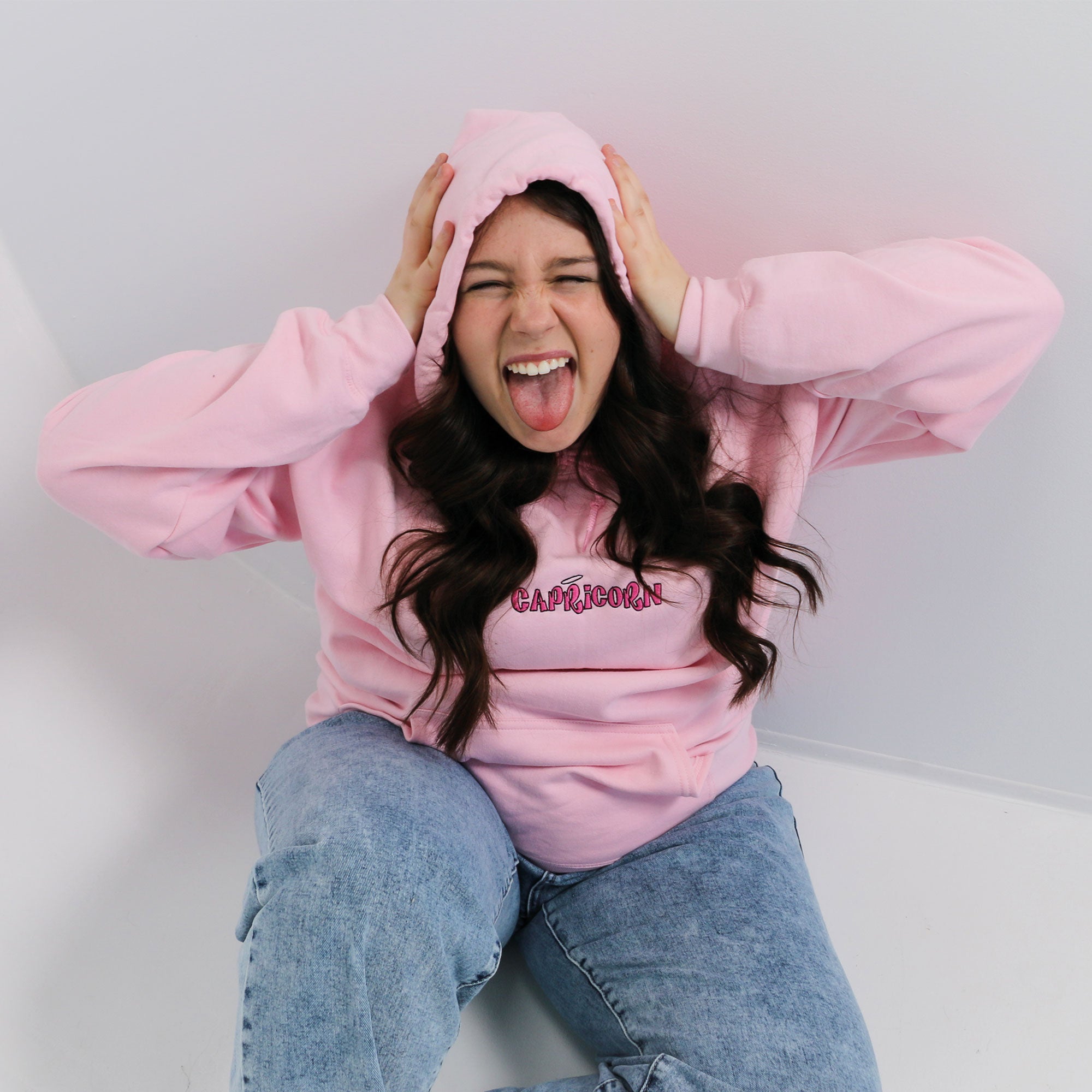 photo lifestyle d'une jeune femme faisant une grimace aux cheveux bruns portant un hoodie rose avec une broderie capricorne personnalisée dans un studio
