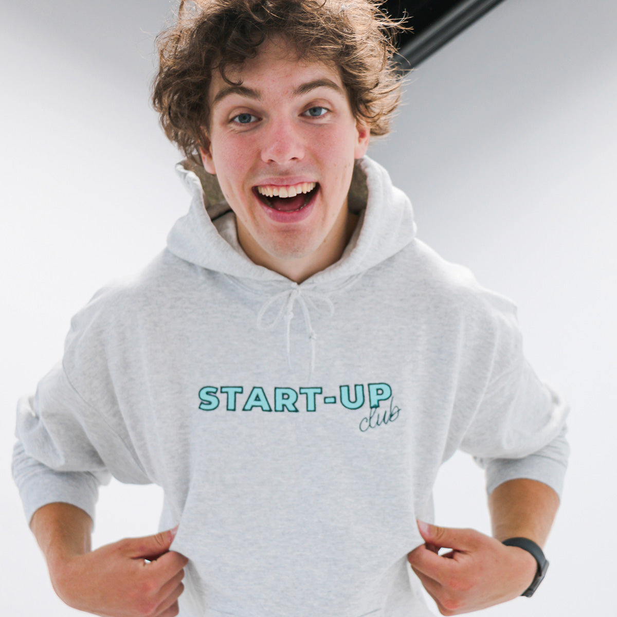 coton ouaté à capuchon gris cendré avec une broderie "start-up club" en turquoise pâle et contour foncé porté par un jeune homme souriant aux cheveux frisés bruns pâles