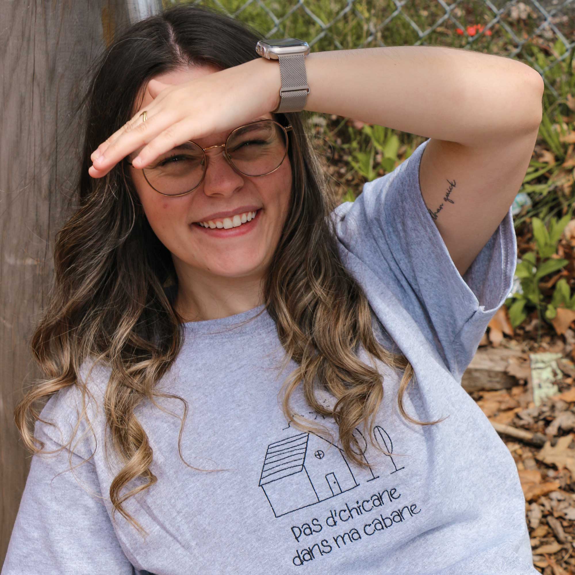 photo lifestyle d'une jeune femme souriante portant un t-shirt gris pâle en manches courtes avec une broderie noire "pas d'chicane dans ma cabane" 