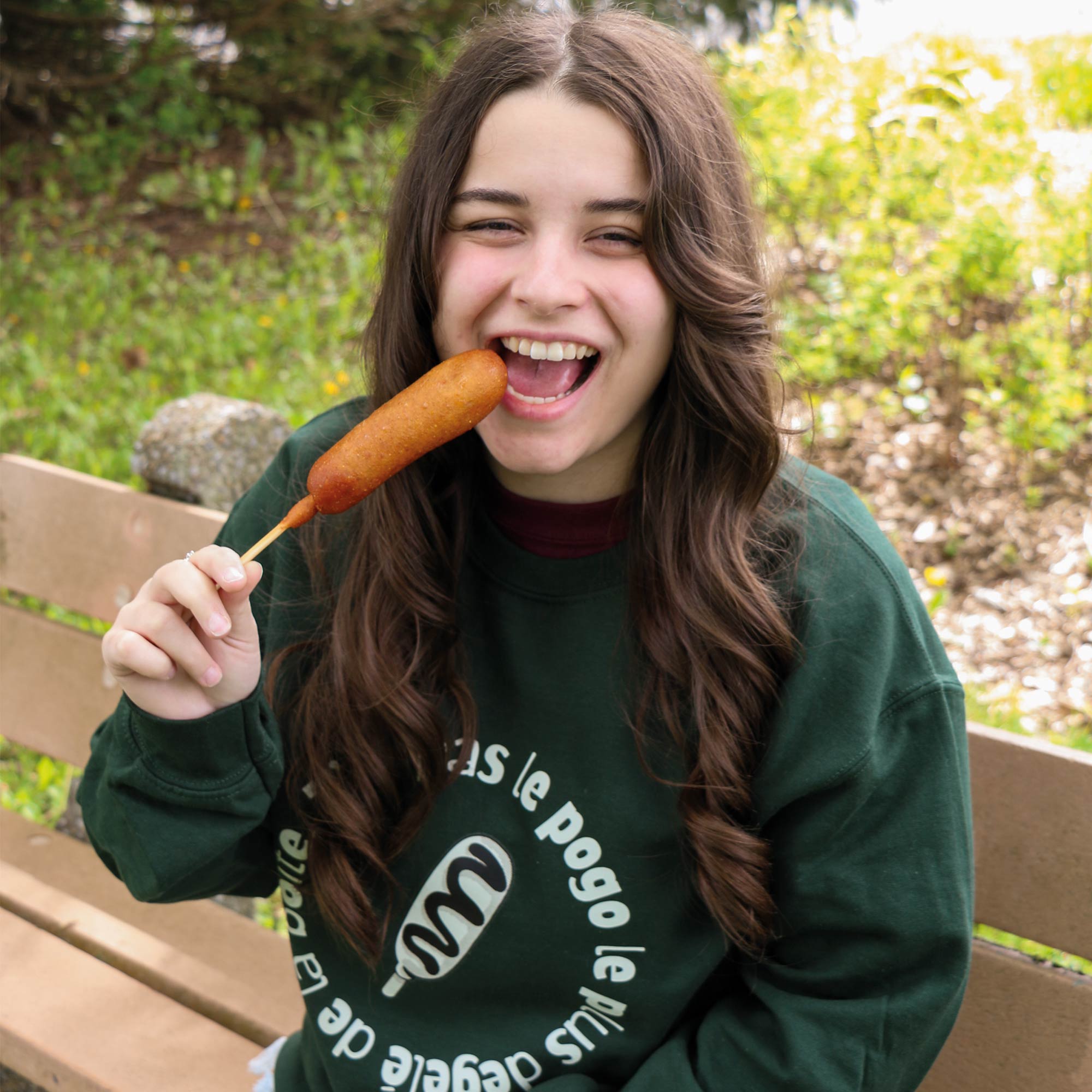 photo lifestyle d'une jeune femme portant un crewneck vert foret avec un imprimé de pogo et l'expression québécoise "t'es pas le pogo le plus dégelé de la boîte". Elle a un pogo dans ses mains et fait semblant de prendre une bouchée