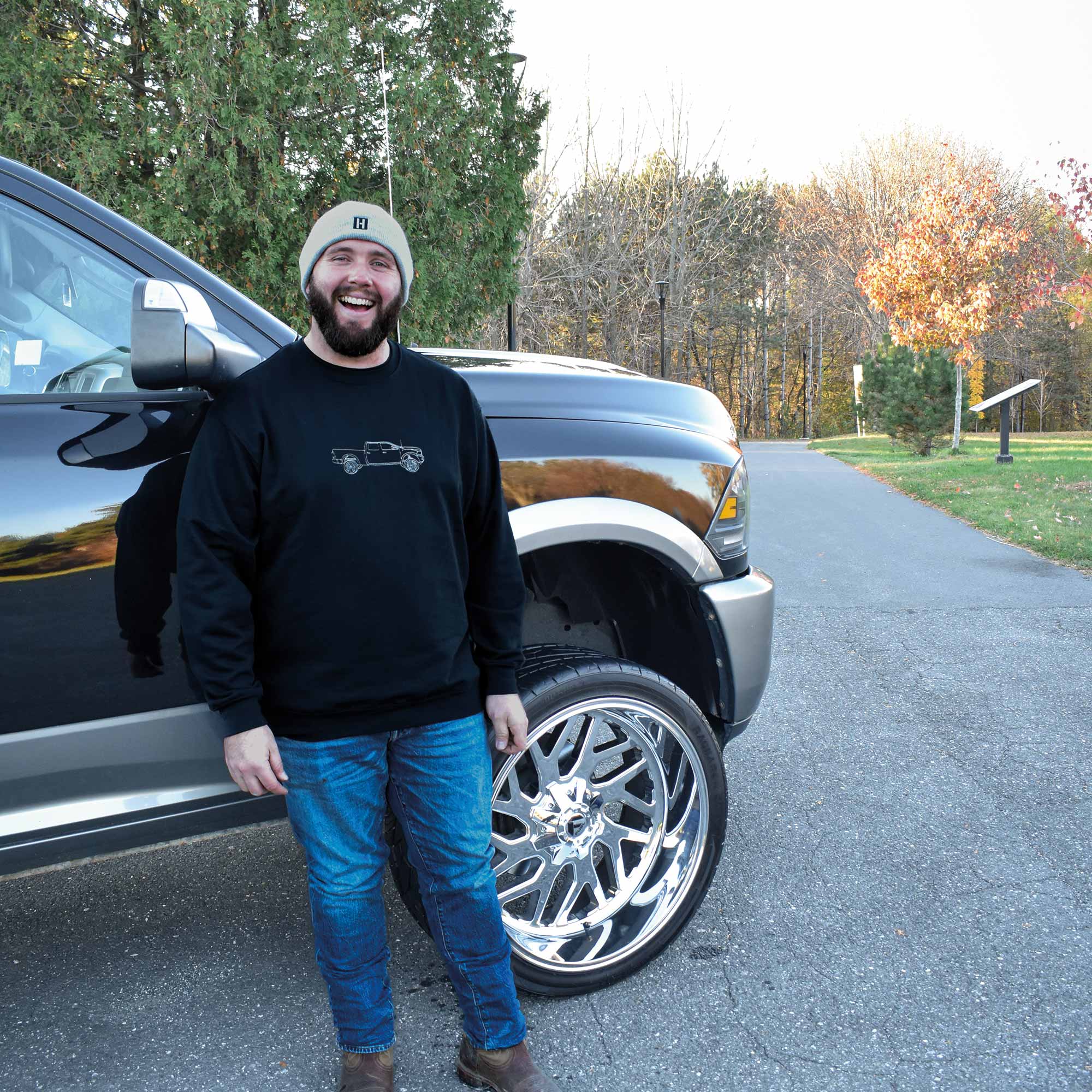 lifestyle homme souriant portant un coton ouaté sans capuchon noir avec son pickup voiture brodée de format silhouette tracée à la main posant devant sa voiture