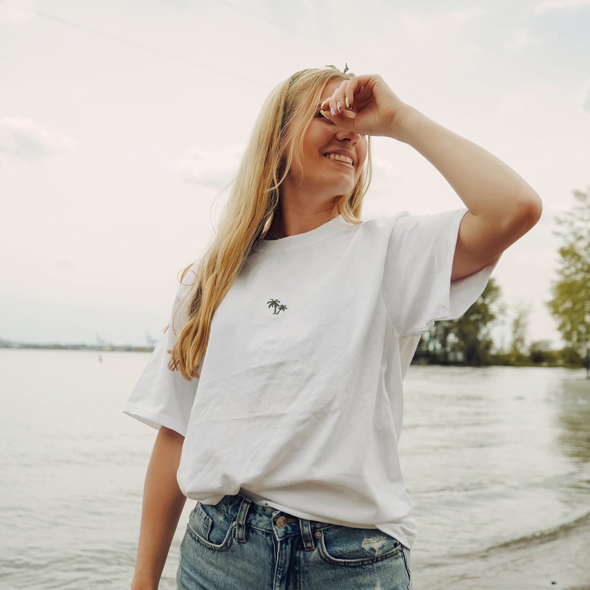 jeune femme blonde souriante sur le bord de la plage portant un t-shirt blanc de palmiers fait par brodame