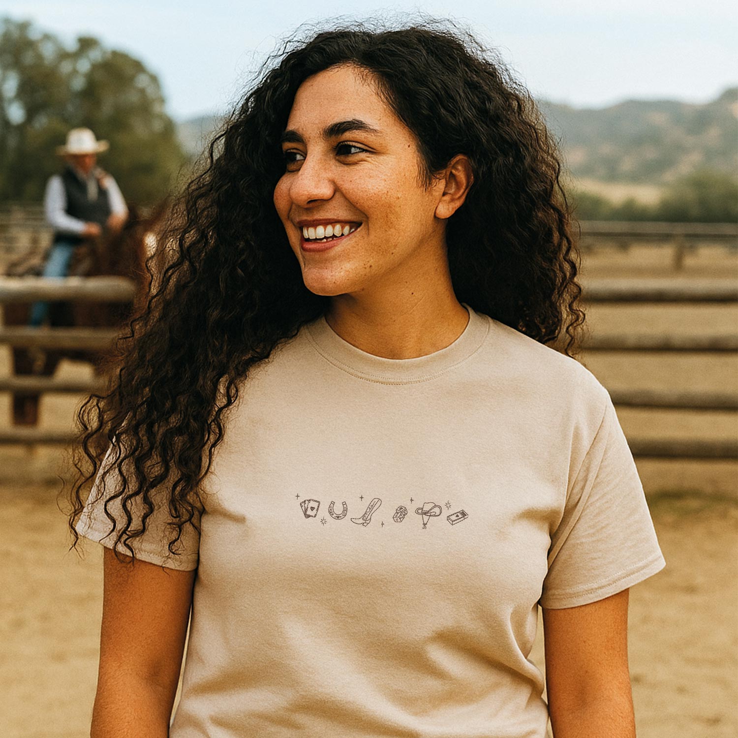 photo lifestyle d'une jeune femme latina aux cheveux bruns frisés portant un t-shirt beige avec une broderie avec des icones western minimalistes brodés en brun. Elle est dans un ranch de chevaux