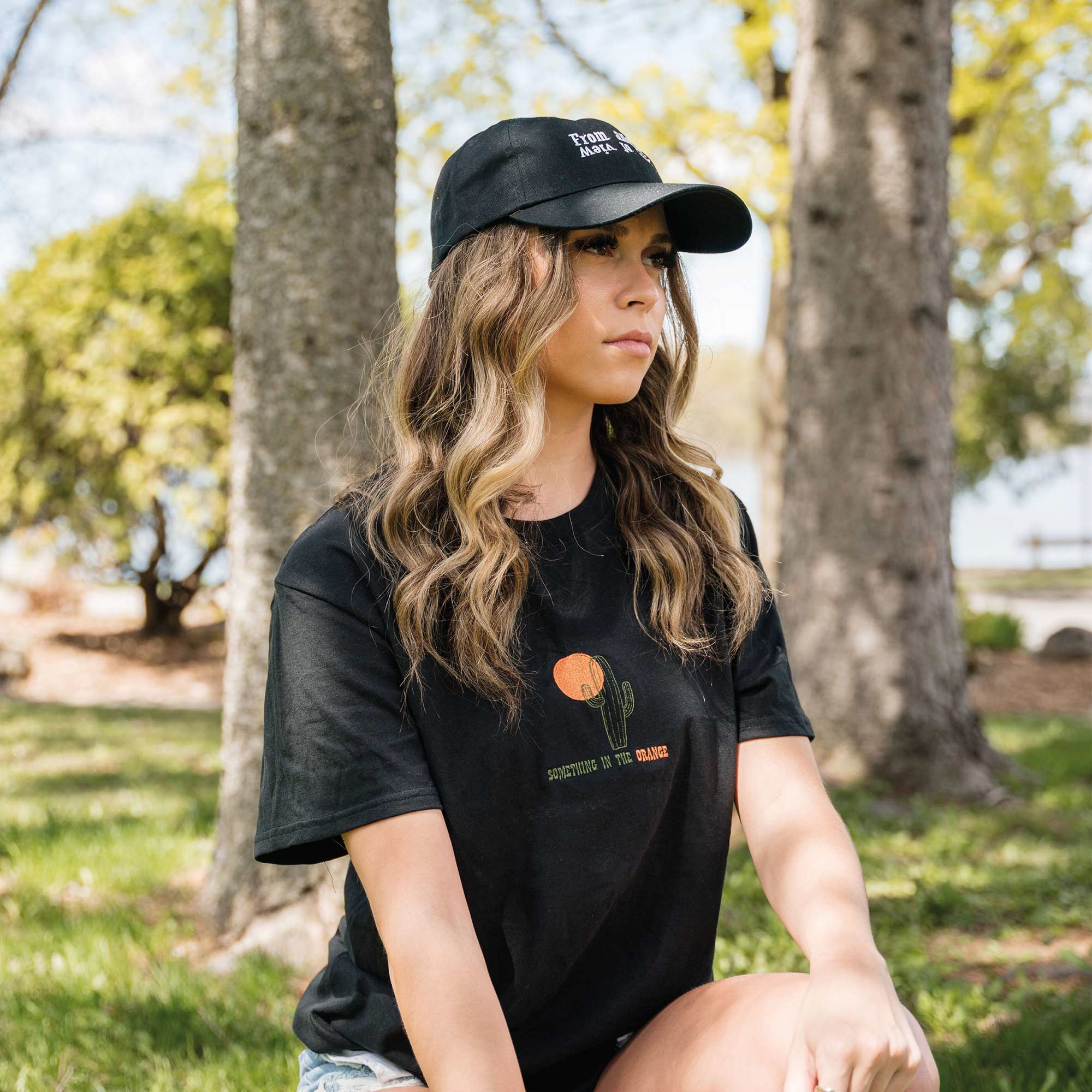 jeune femme portant un t-shirt noir avec broderie soleil et cactus something in the orange country avec une casquette noir POV