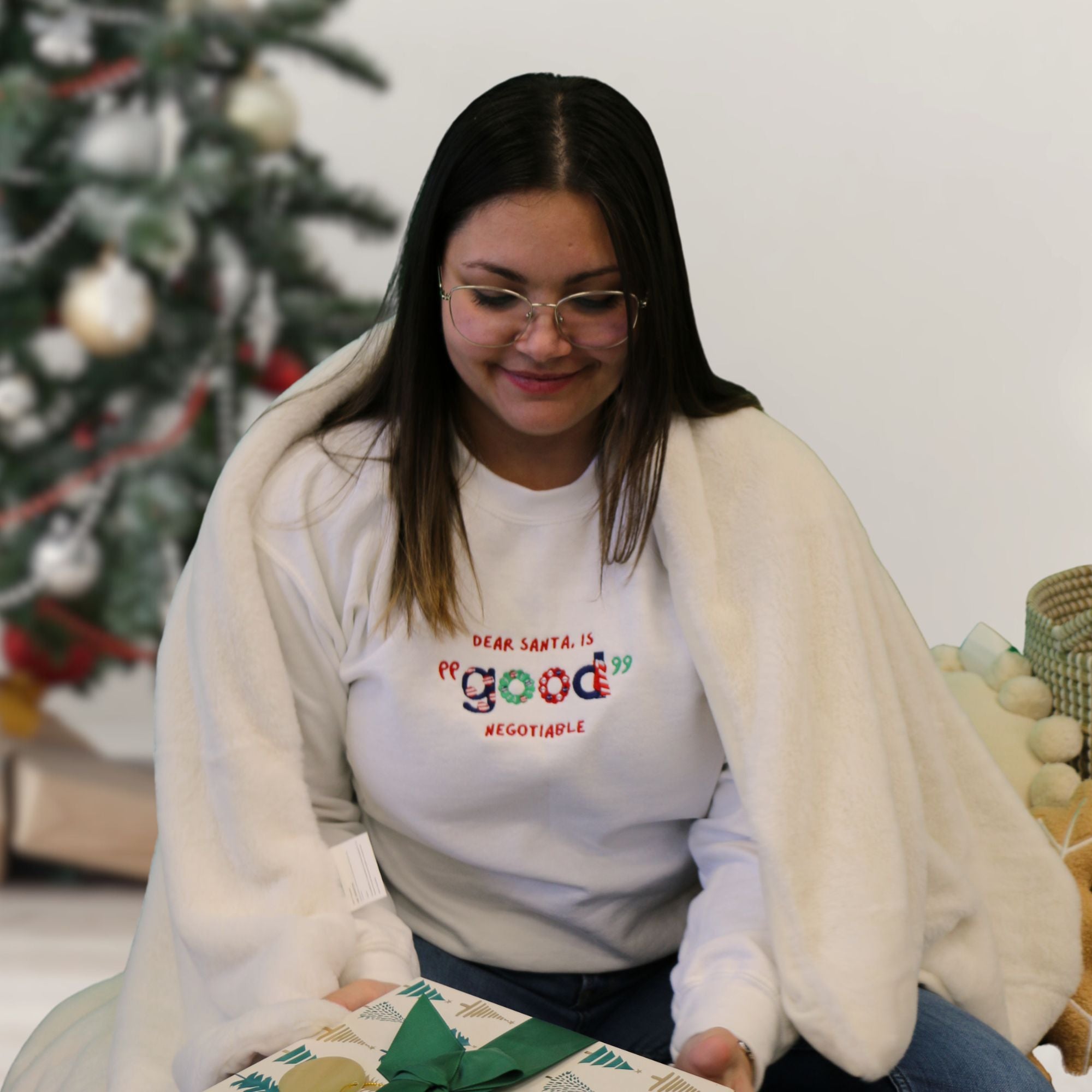 photo d'une jeune femme portant un crewneck brodé "dear santa, is good negociable" blanc devant un sapin de noel