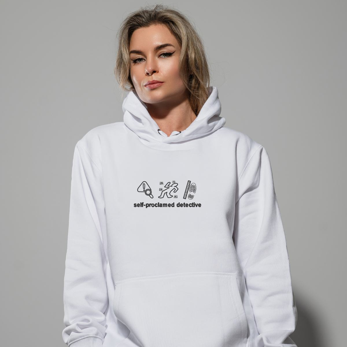 photo dans un studio d'une femme blonde portant un hoodie blanc avec une broderie "self-proclamed detective" au centre, sur fond gris