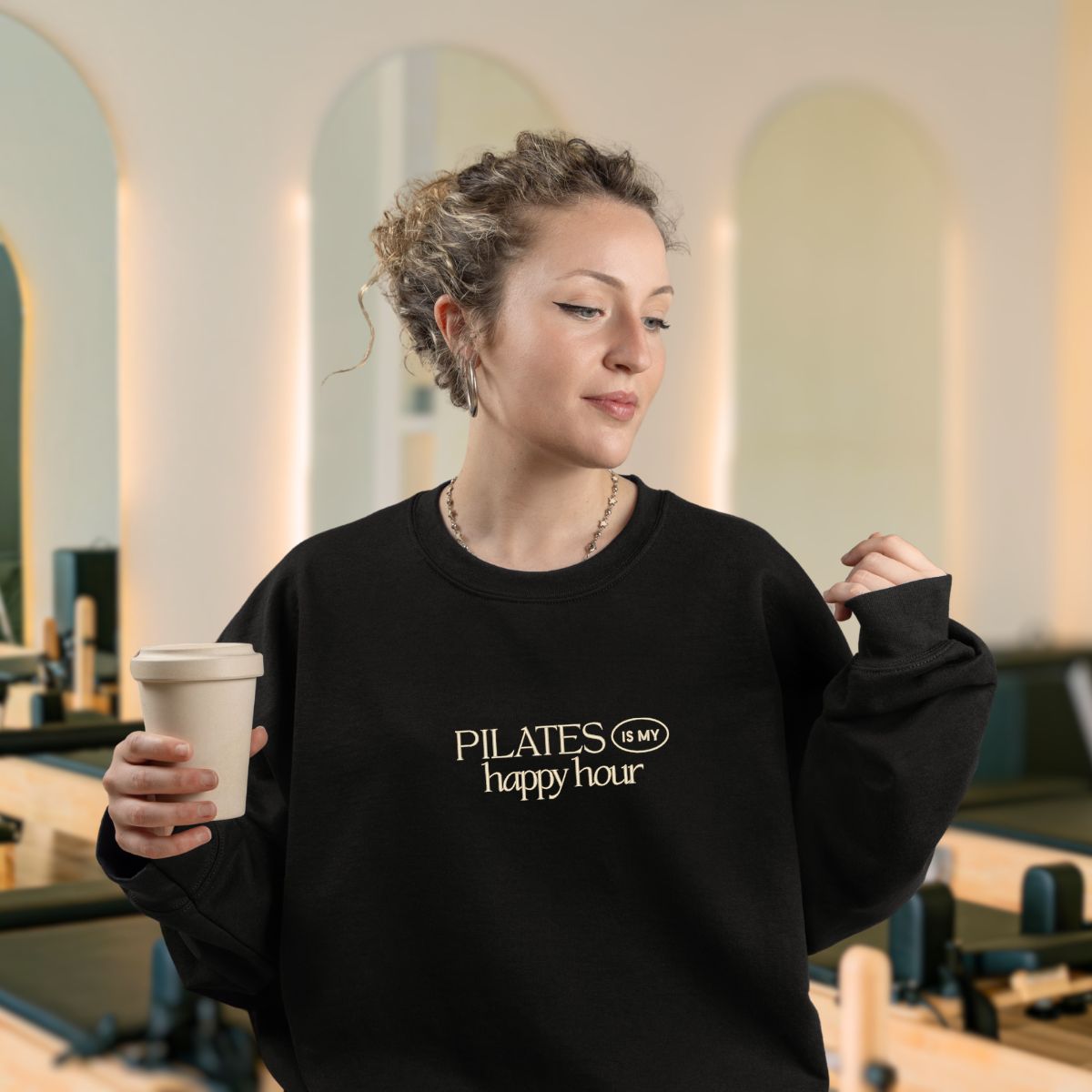 photo lifestyle d'une prof de pilates au cheveux longs bouclés qui est dans son studio buvant son matcha en portant son crewneck noir avec une broderie "pilates is my happy hour" en crème.