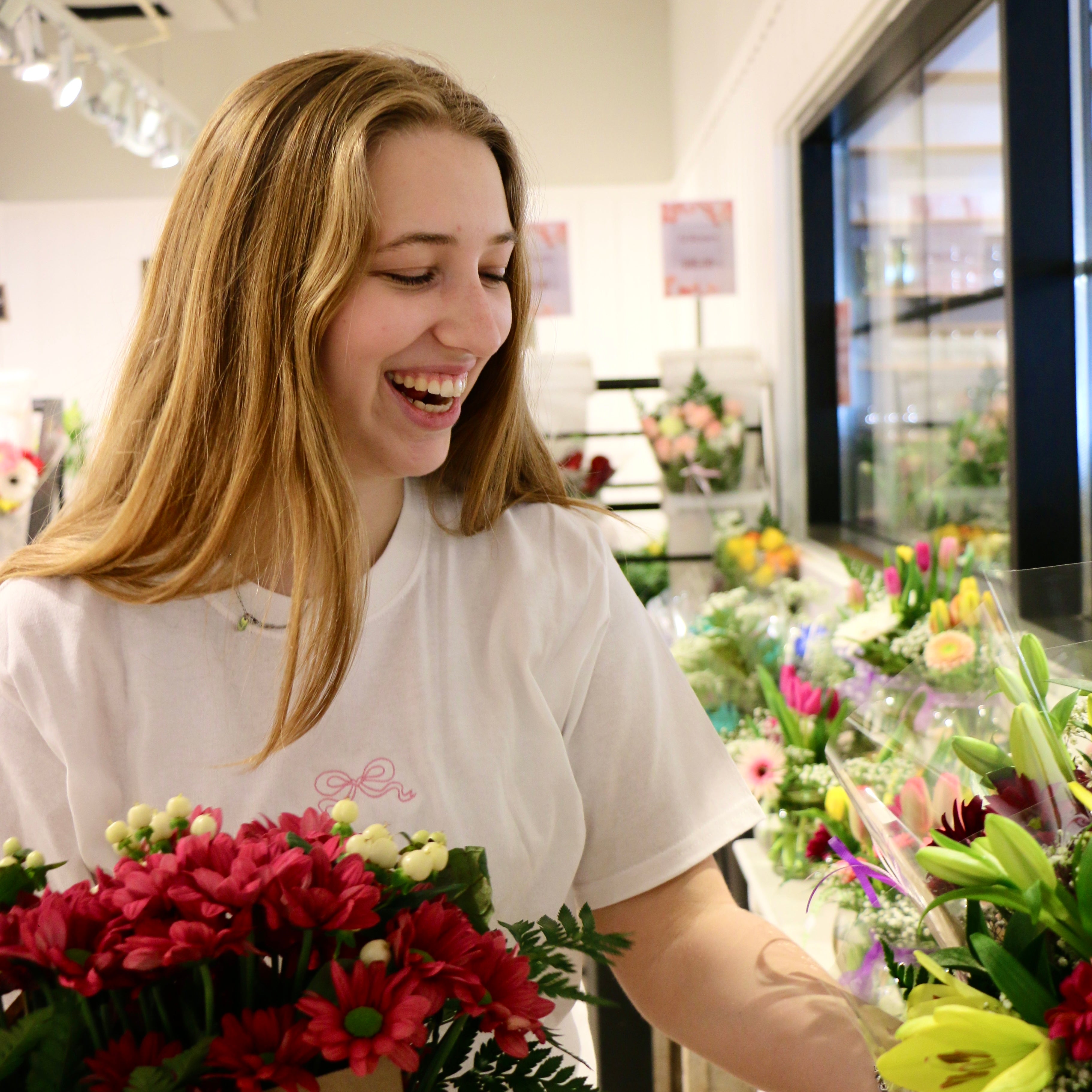 photo d'une jeune femme portant un t-shirt blanc ayant une petite boucle rose brodée au centre. photo prise chez un fleuriste