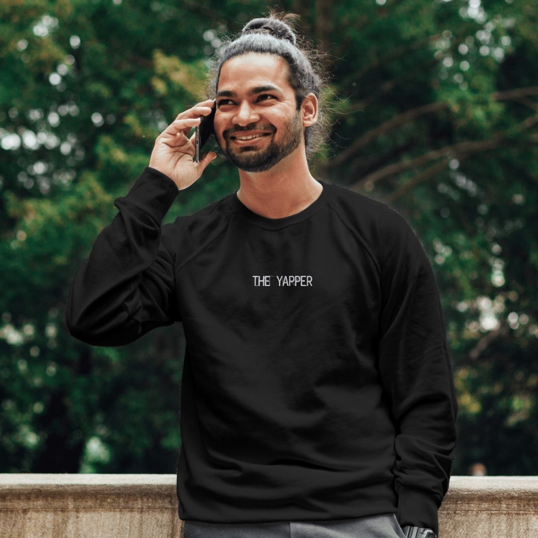 coton ouaté à col rond noir avec broderie "the yapper" en blanc porté par un homme d'origine indienne qui parle au téléphone