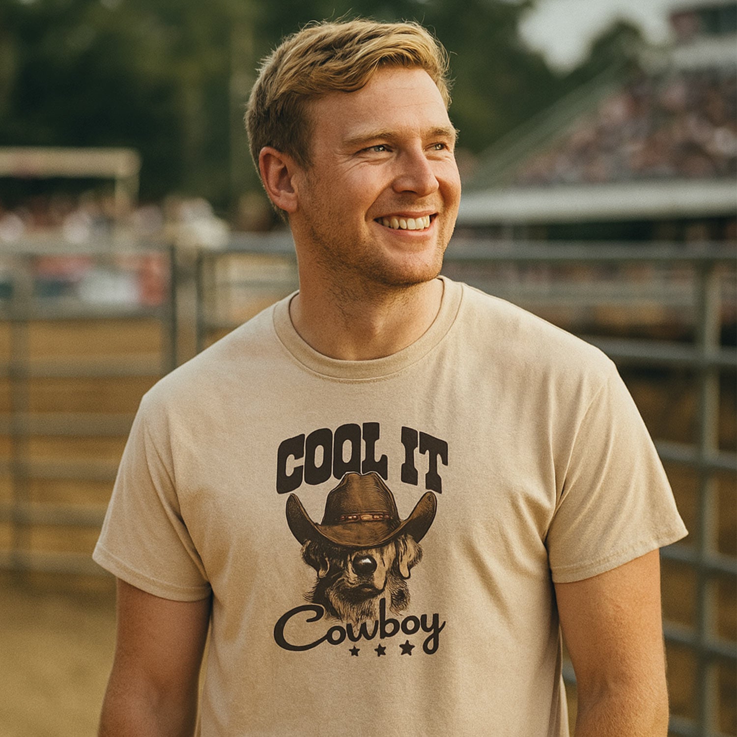 jeune homme aux cheveux blonds dans un décor de rodéo portant un t-shirt beige à manches courtes imprimé avec un golden retriver portant un chapeau de cowboy et les mots "cool it cowboy" autour